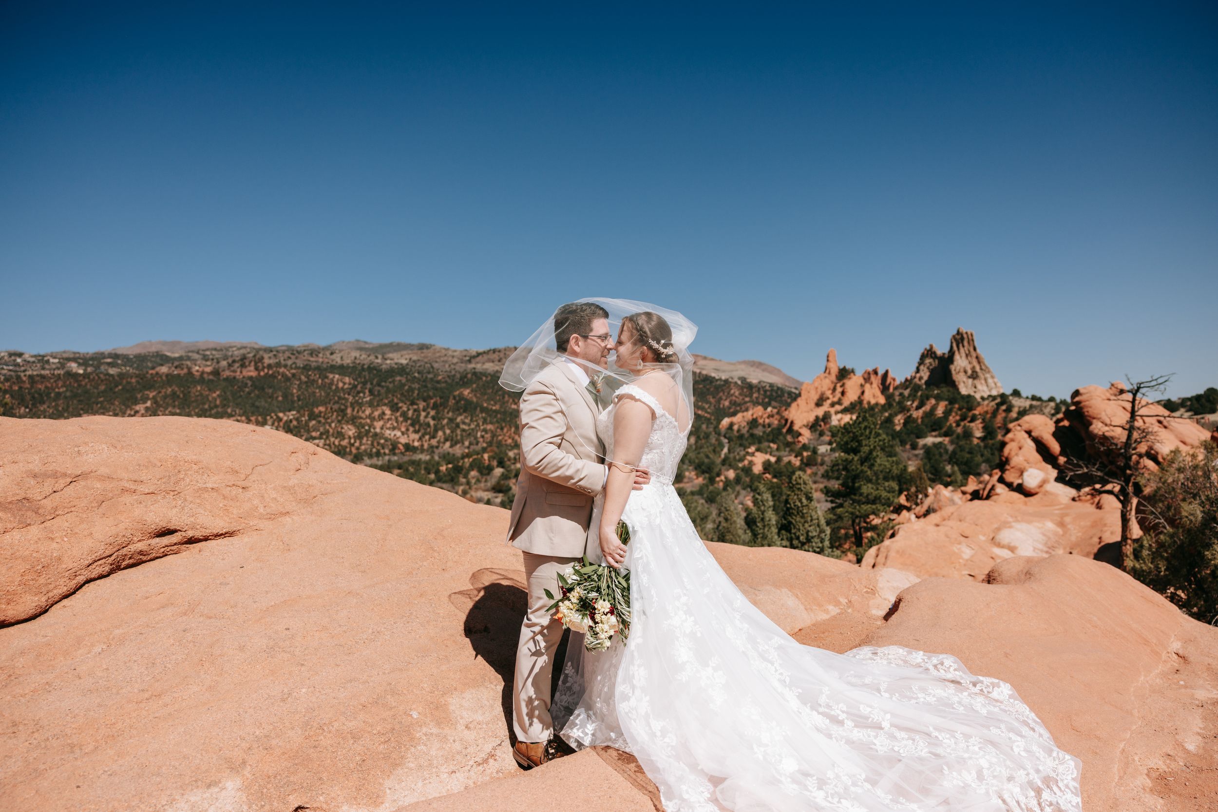 A scenic wedding photo captured on red rock formations in Garden of the Gods with blue sky and mountains in background.