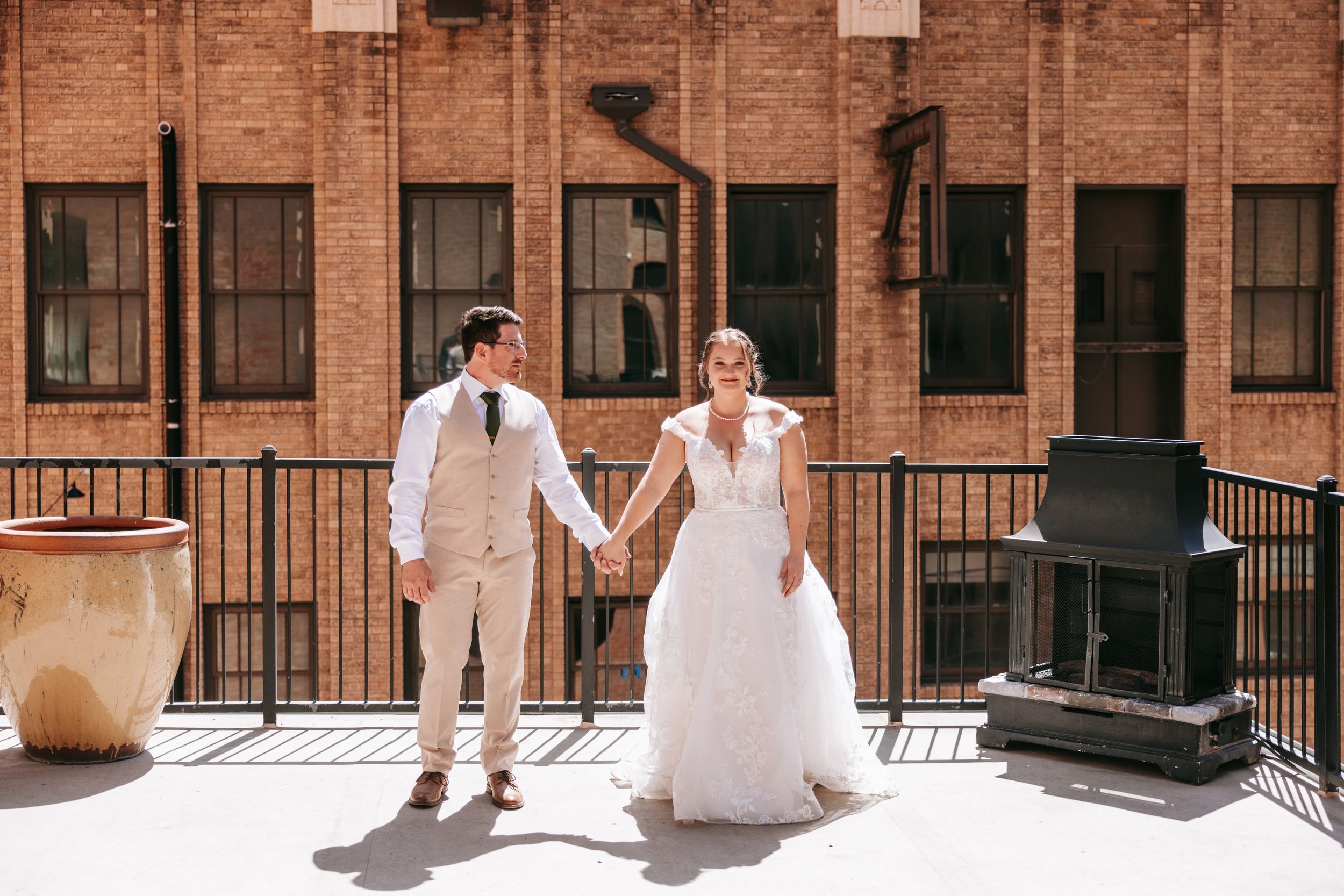 A joyful wedding couple holds hands while standing on an outdoor terrace with brick walls and railings in the background.