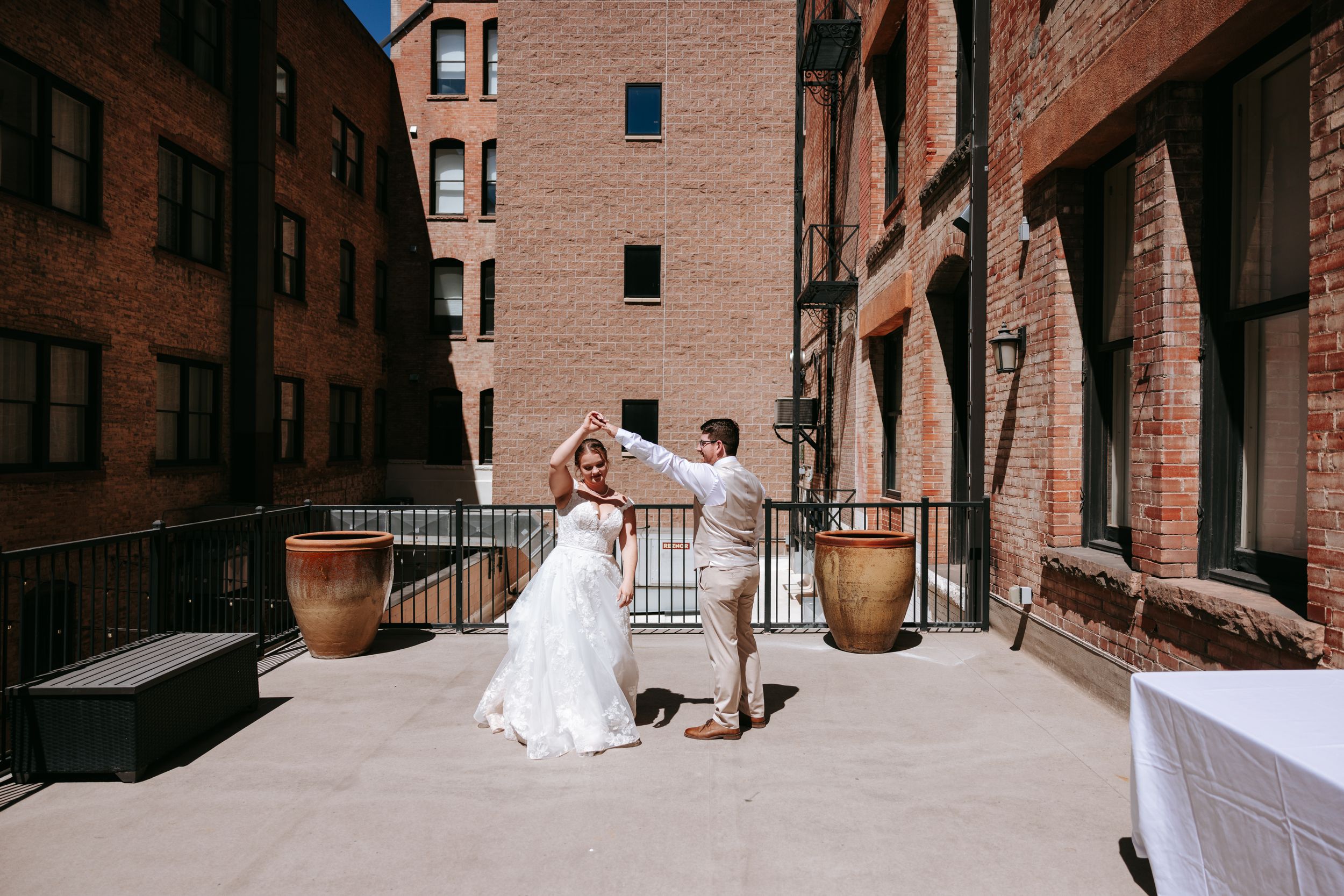 A couple in wedding attire dances on a rooftop terrace surrounded by brick buildings and potted plants.