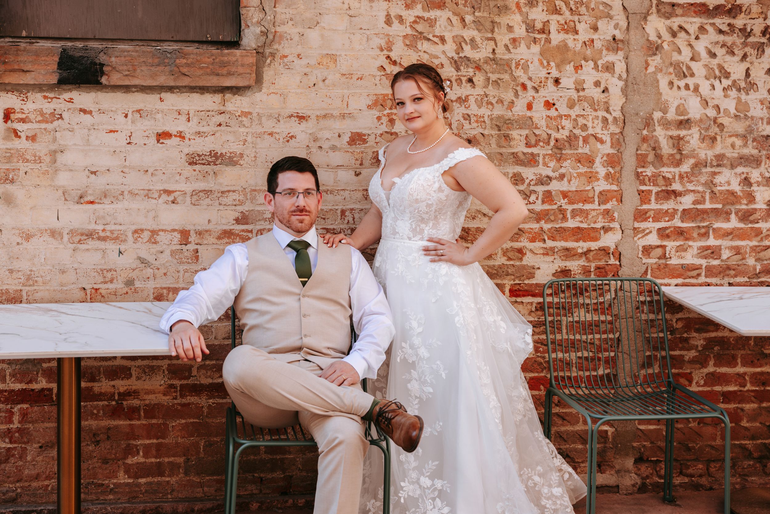 A bride in white lace dress and groom in beige suit pose against a rustic brick wall.