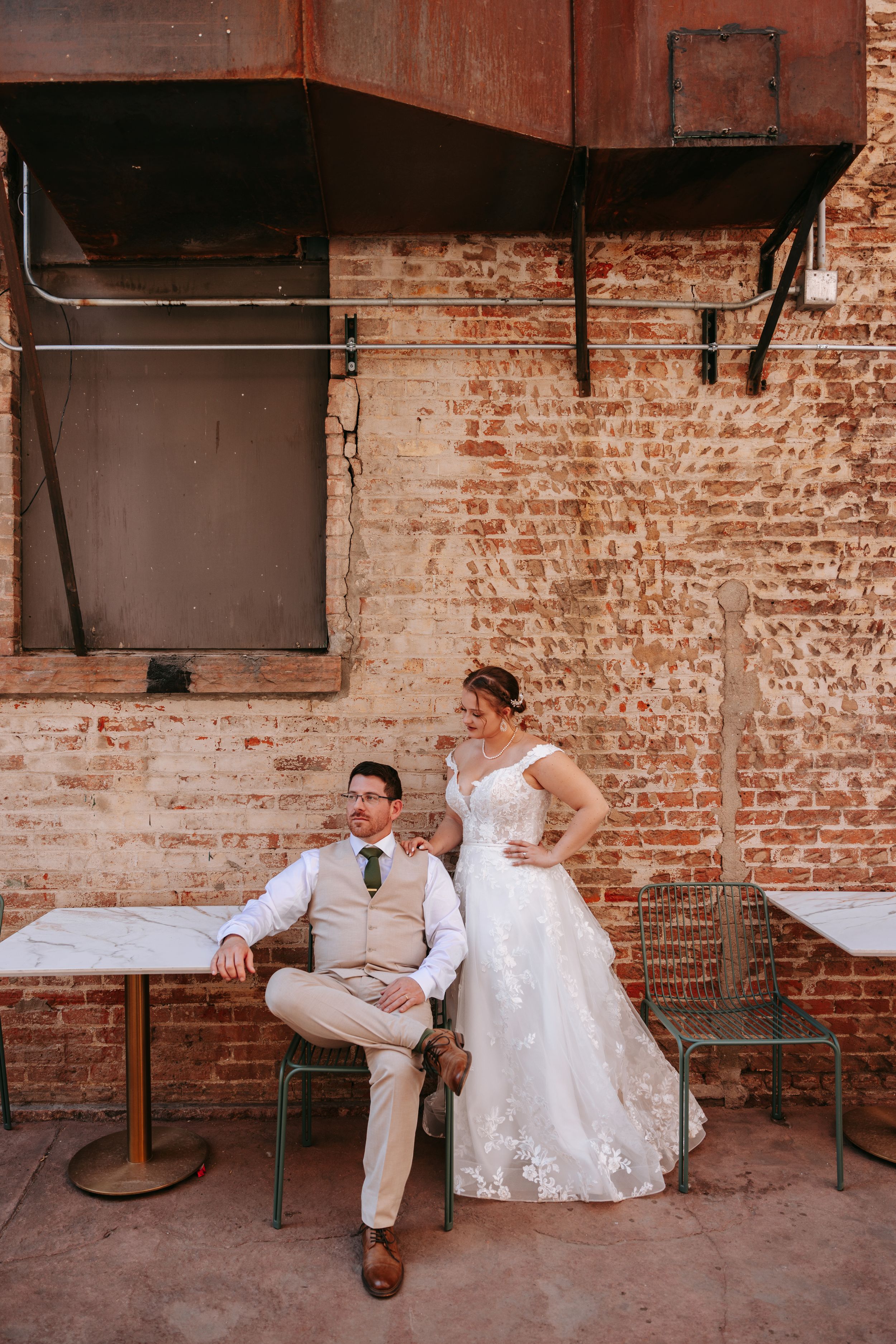 Couple in wedding attire poses by rustic brick wall with vintage industrial decor.