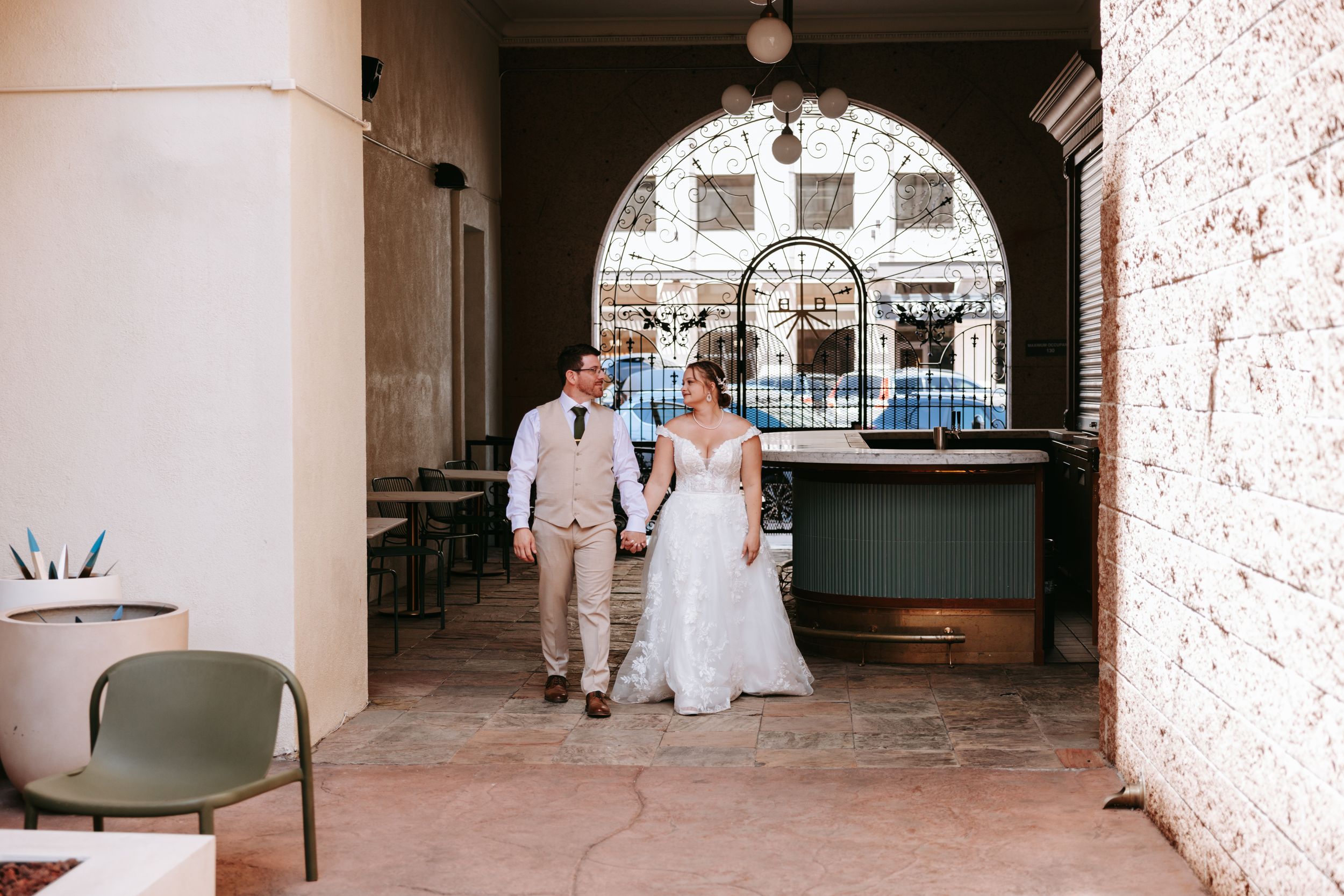 A couple in wedding attire walks through an arched brick passageway with outdoor seating and retro decor.