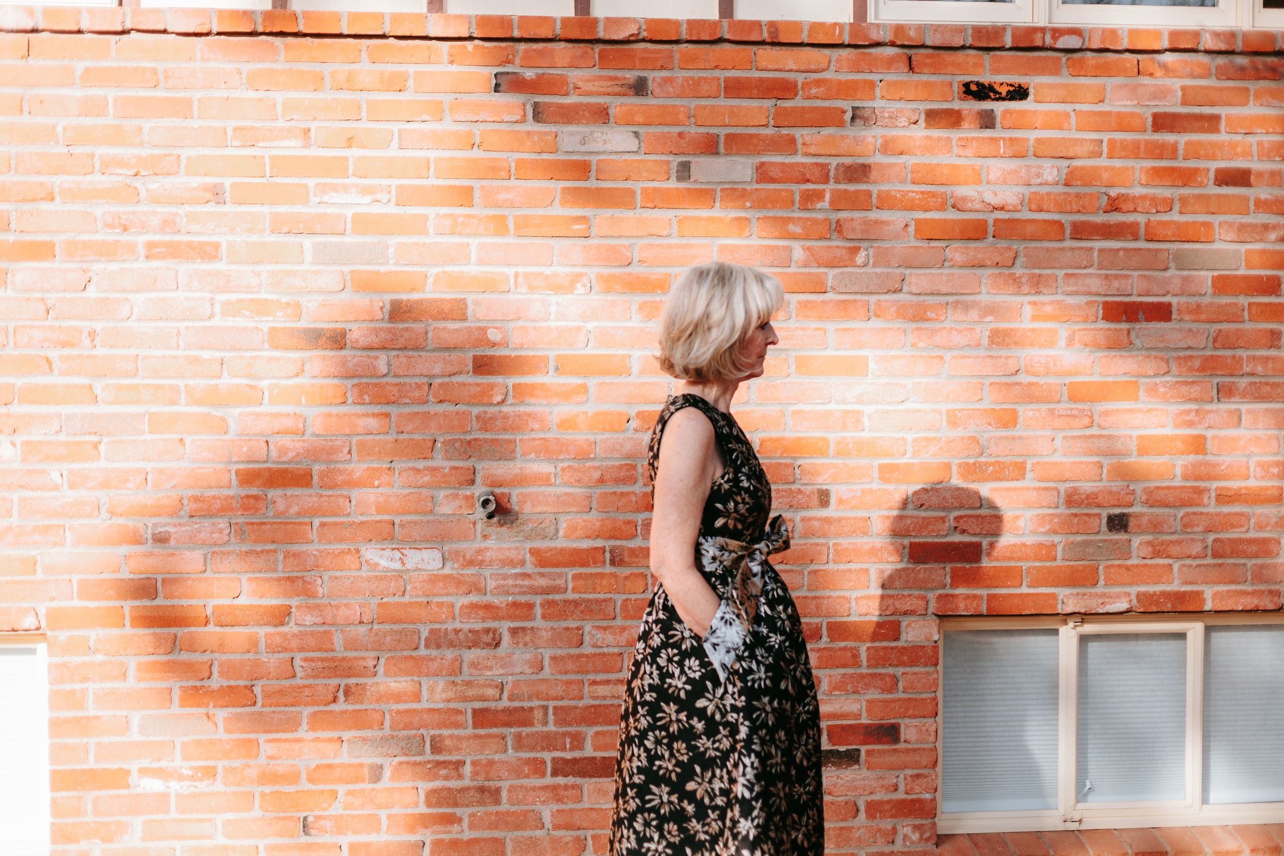 Someone in a floral dress poses against a sun-lit brick wall during a bright summer day.