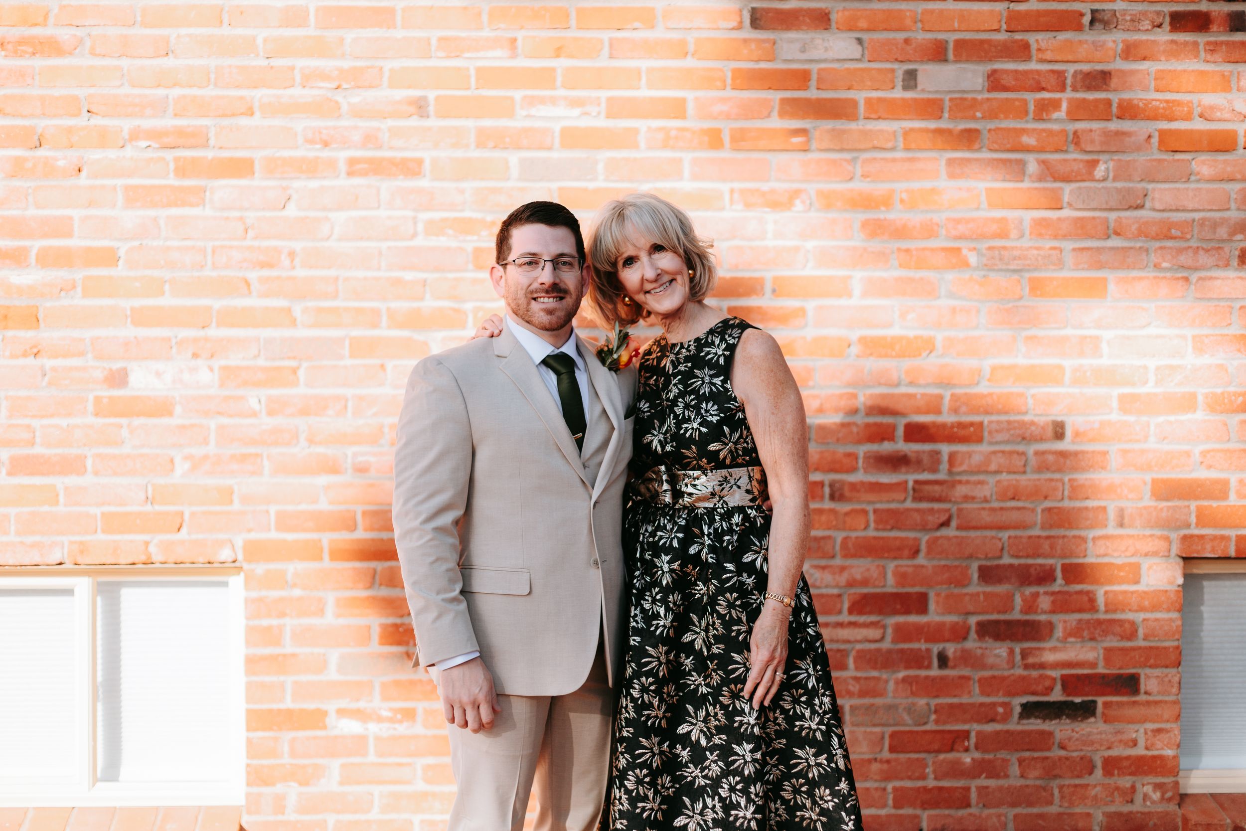 A couple stands against a rustic brick wall, dressed formally with the person in a tan blazer and another in floral dress.