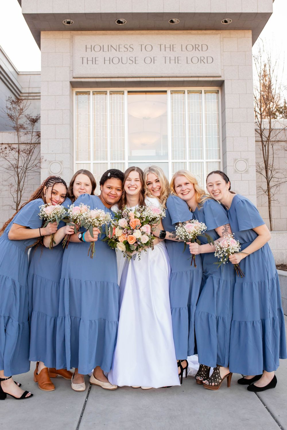 A bridal party posing outside a temple entrance wearing dusty blue dresses and holding floral bouquets.