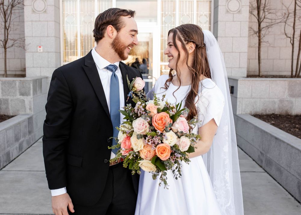 Joyful newlyweds share a tender moment outside stone building with bride holding peach and coral floral bouquet.
