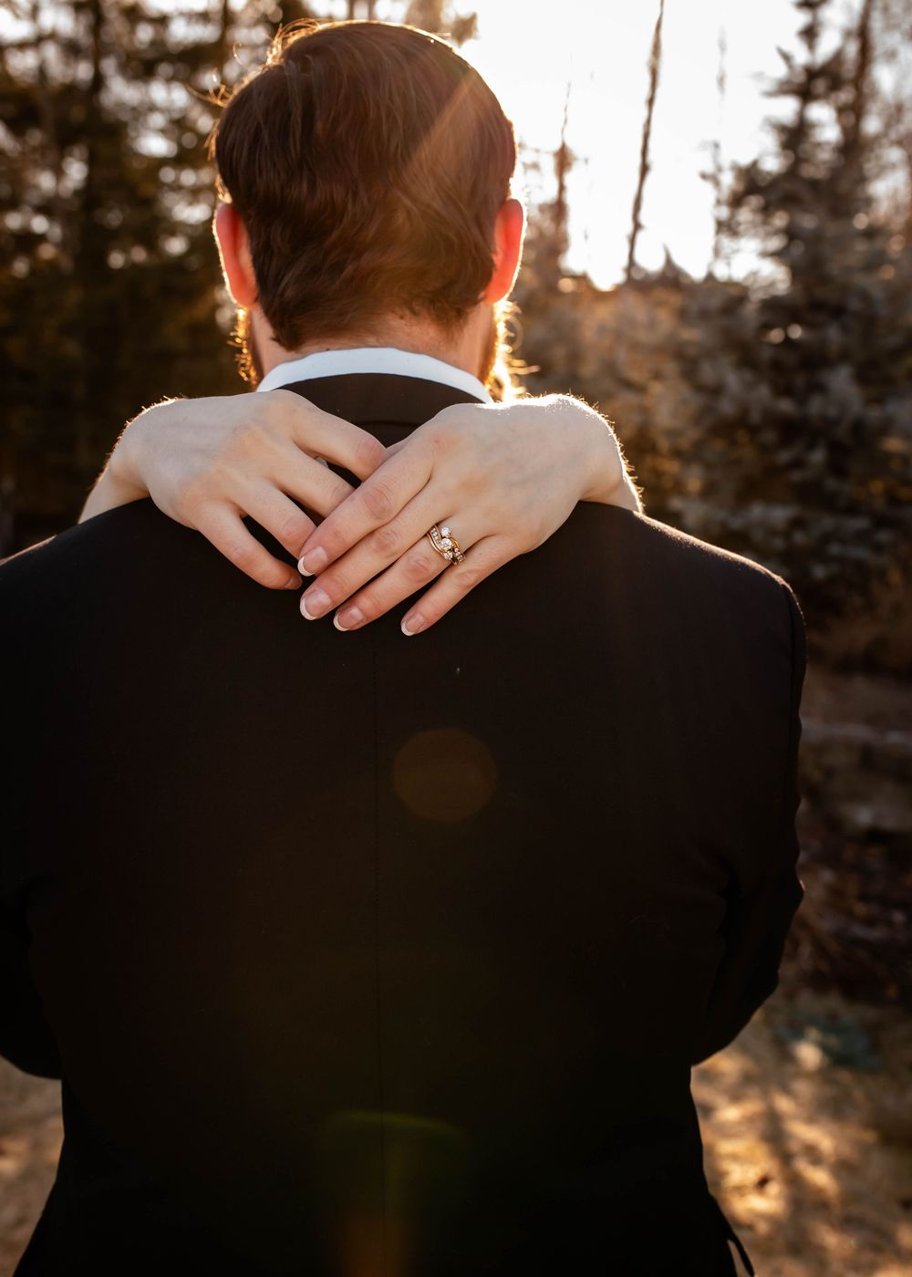 Bride's hand with engagement ring rests on groom's shoulder during golden hour sunset with trees in background.