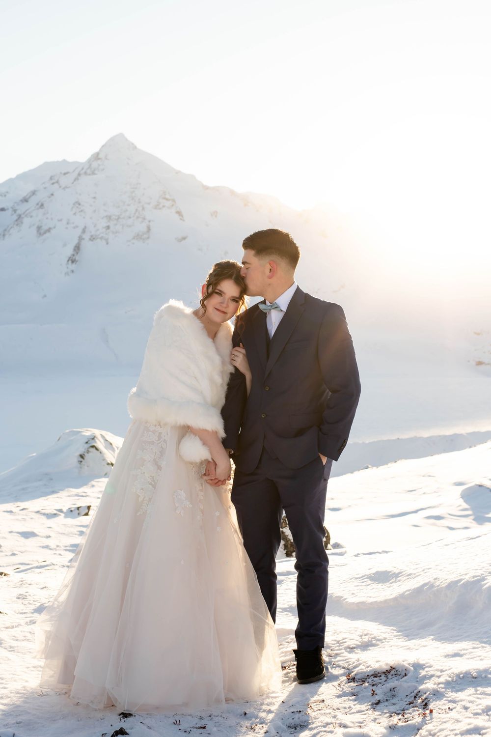 Romantic winter wedding photos captured in snowy mountains with dramatic backlighting at sunset.