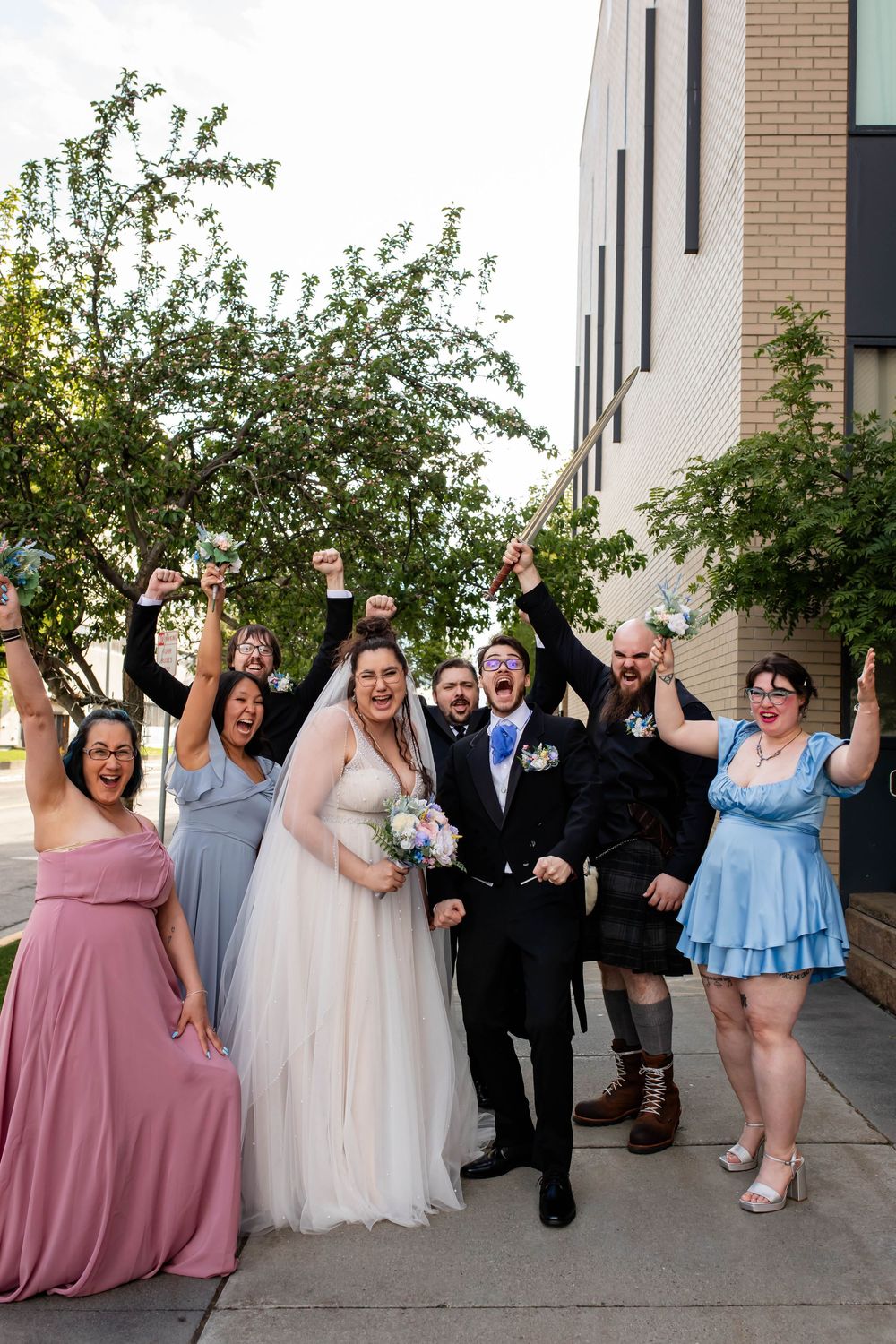 Wedding party in pink, gray and blue dresses celebrating joyfully outside a brick and white building.