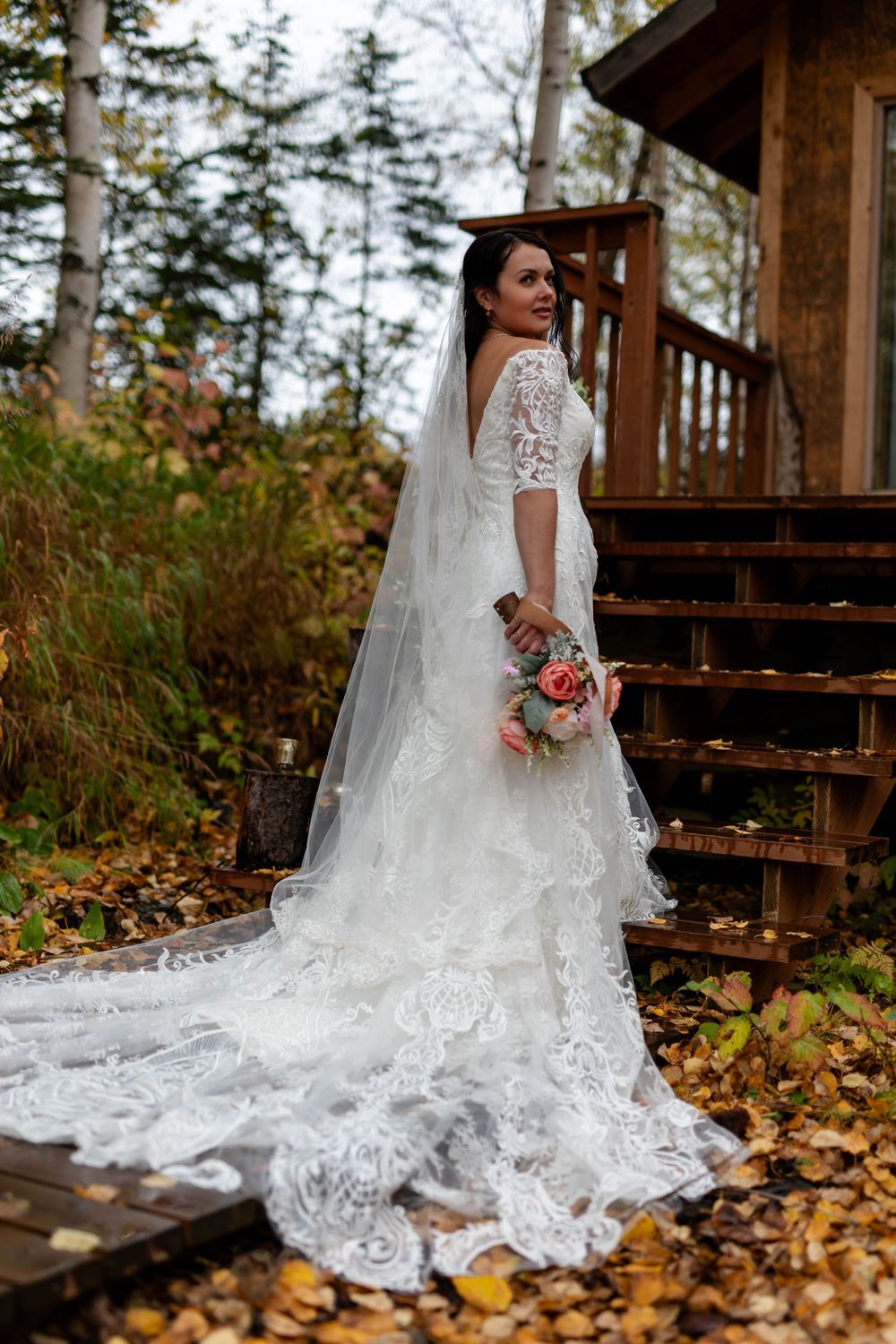 Elegant bride in white lace gown with flowing train poses on rustic wooden steps covered in fallen autumn leaves.
