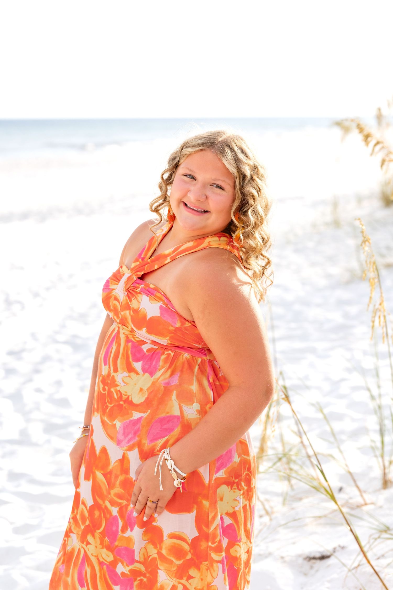 Person in vibrant orange floral maxi dress poses on white sand beach with sea grasses and ocean in background.