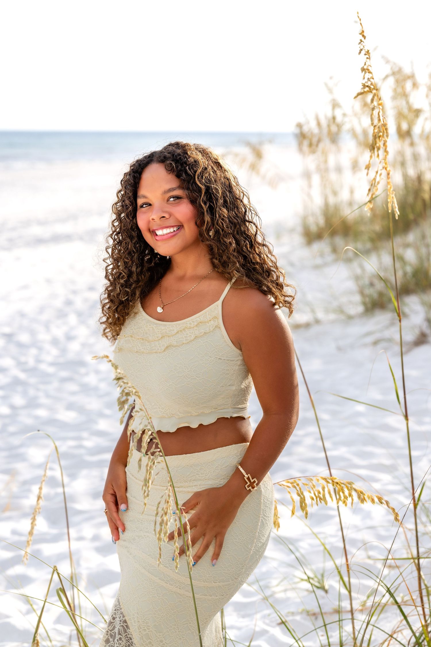 A beachside portrait featuring beautiful curly hair and cream-colored outfit with sea oats and ocean waves in background.