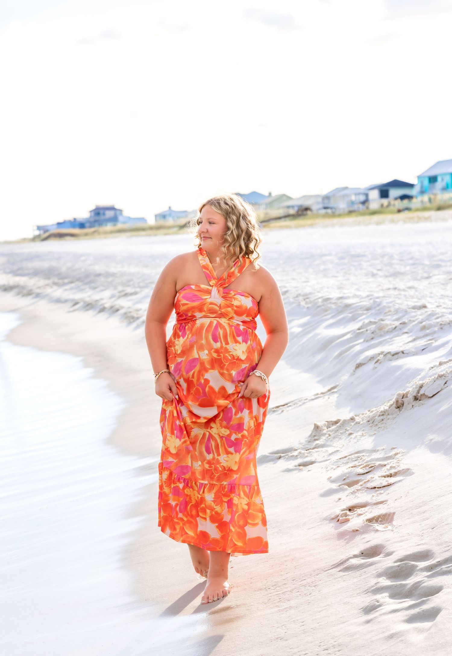 Person in a vibrant orange floral maxi dress walks barefoot along a sandy beach at sunset with coastal homes in background.