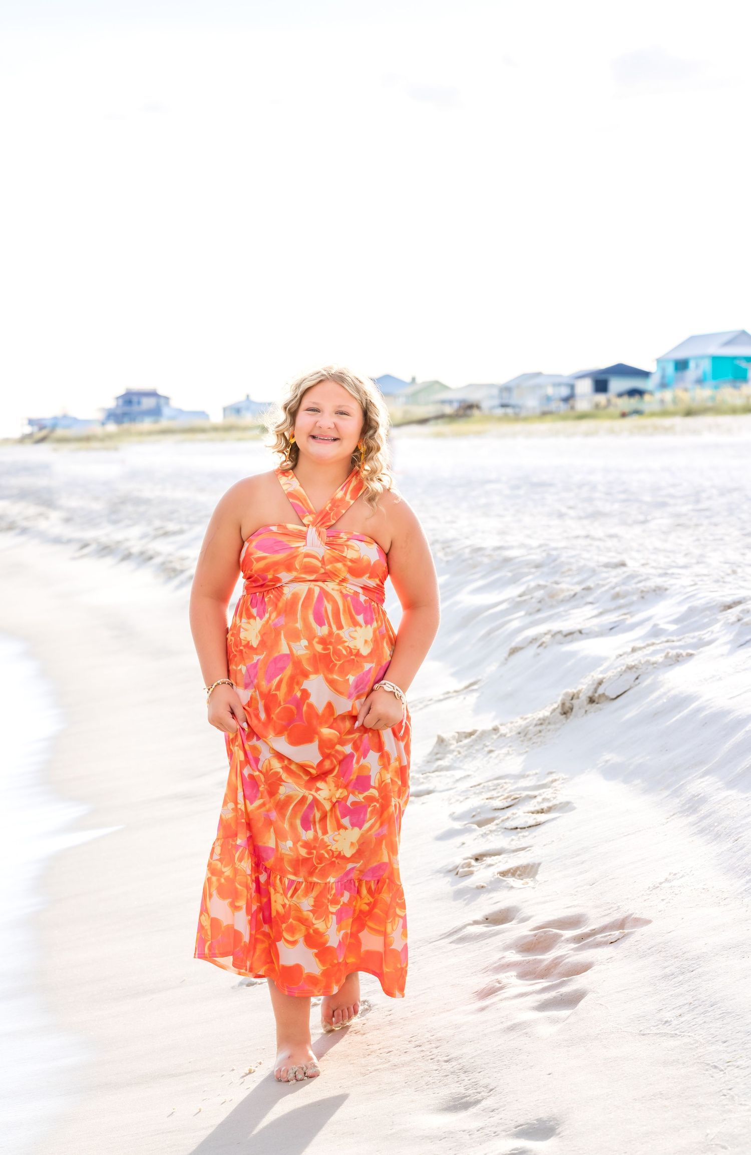 Person wearing coral floral maxi dress walks along shoreline with beach houses visible in background.
