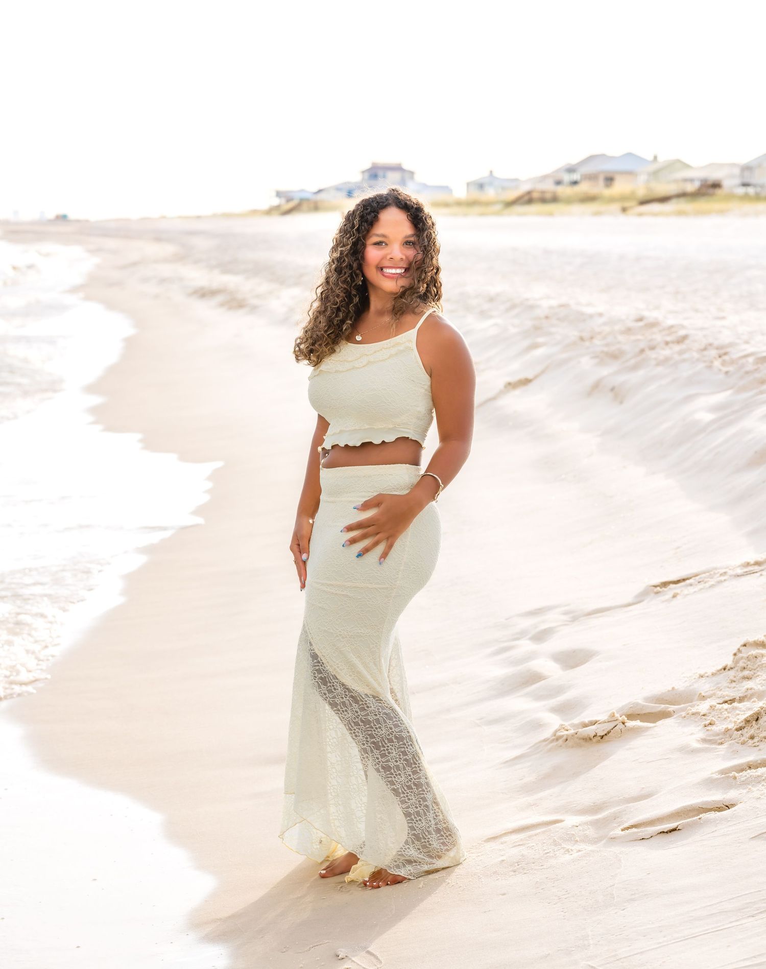 A person poses confidently in a white two-piece outfit while standing on a sandy beach with dunes in the background.