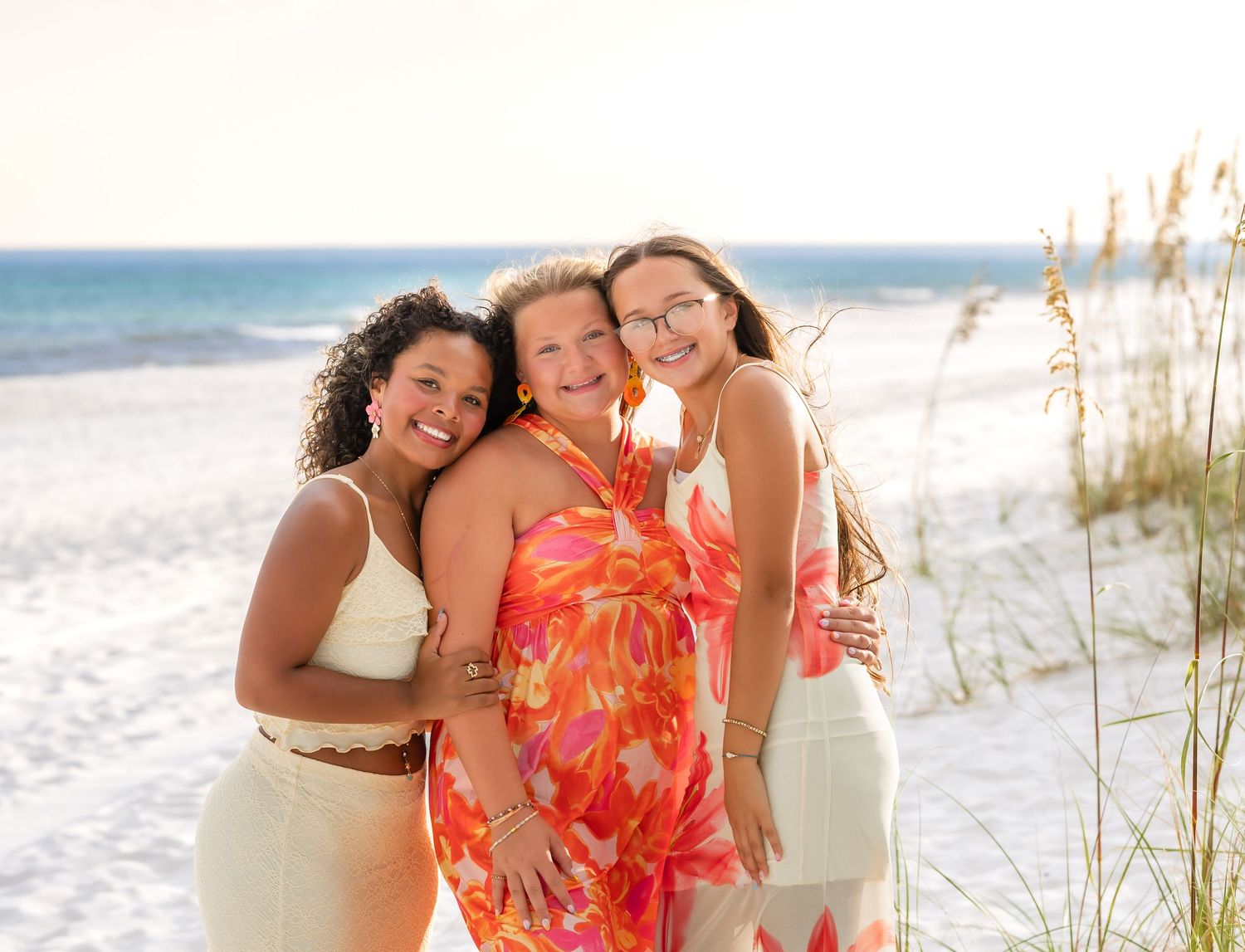 Three friends in floral and white dresses embrace while smiling on a sunny beach with sea grass and ocean waves.