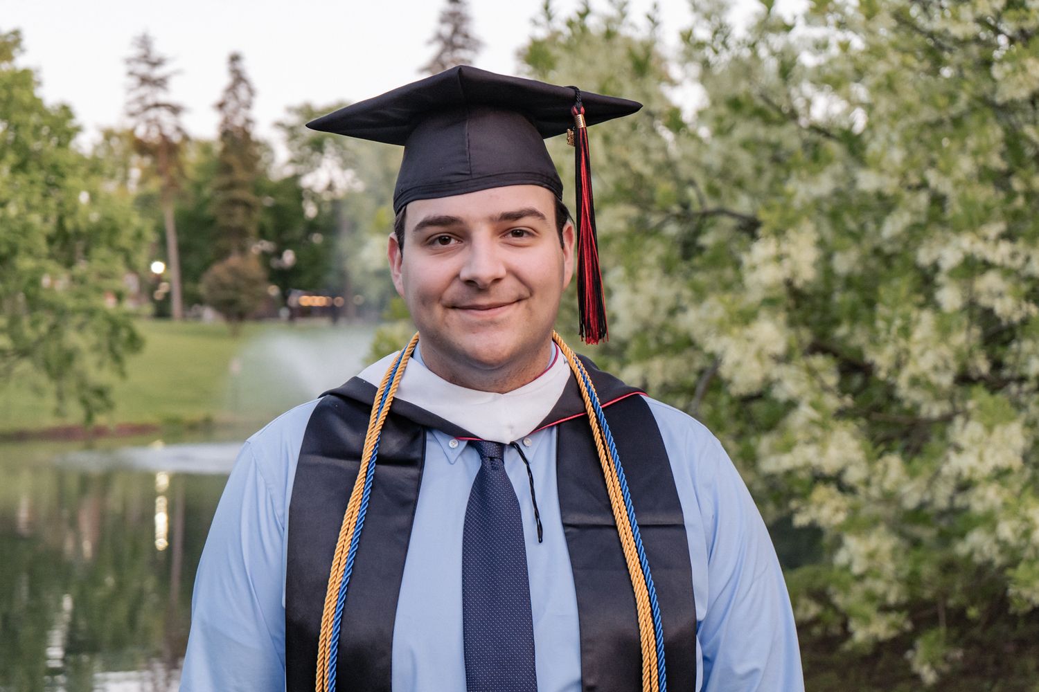 Graduate in academic regalia smiles for photos outdoors with trees and lake in background.