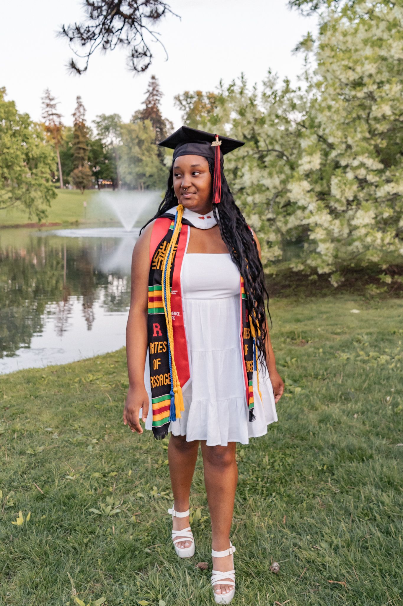 Graduate wearing cap, gown and colorful stole poses on grassy lawn near water.