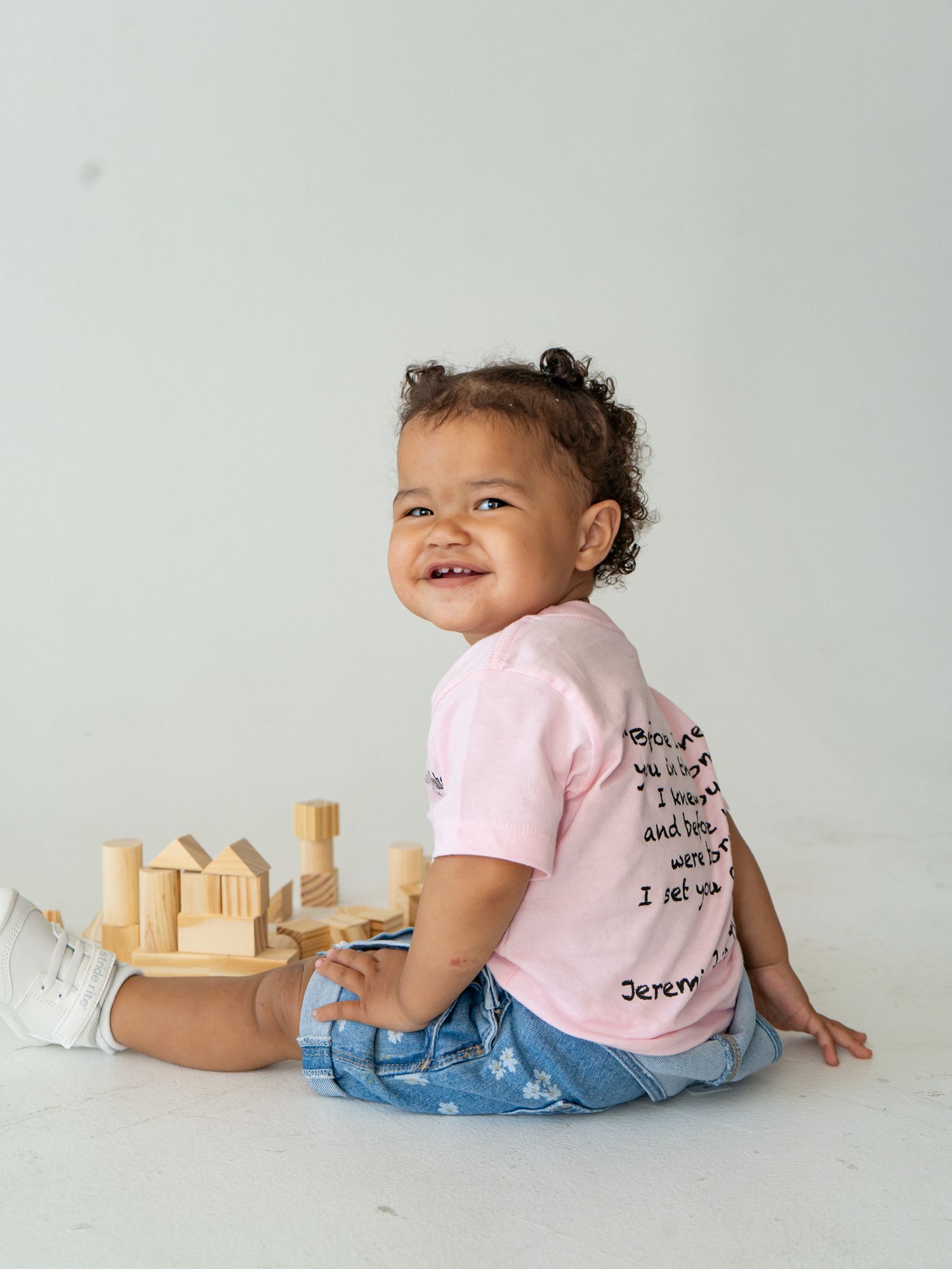 Smiling toddler in pink scripture tee and jeans playing with wooden blocks on white background.