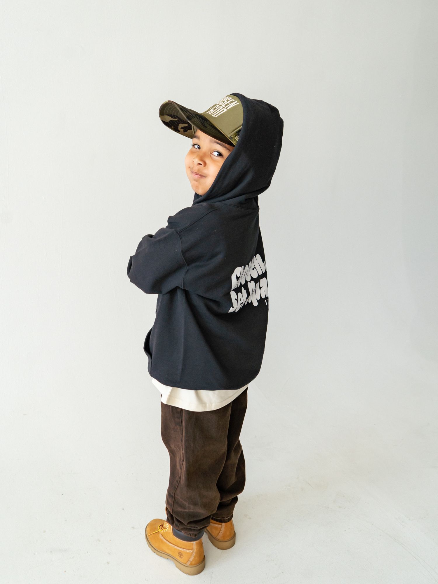 Young boy in navy hoodie, camo cap, brown pants, and tan boots poses with arms crossed against white background.