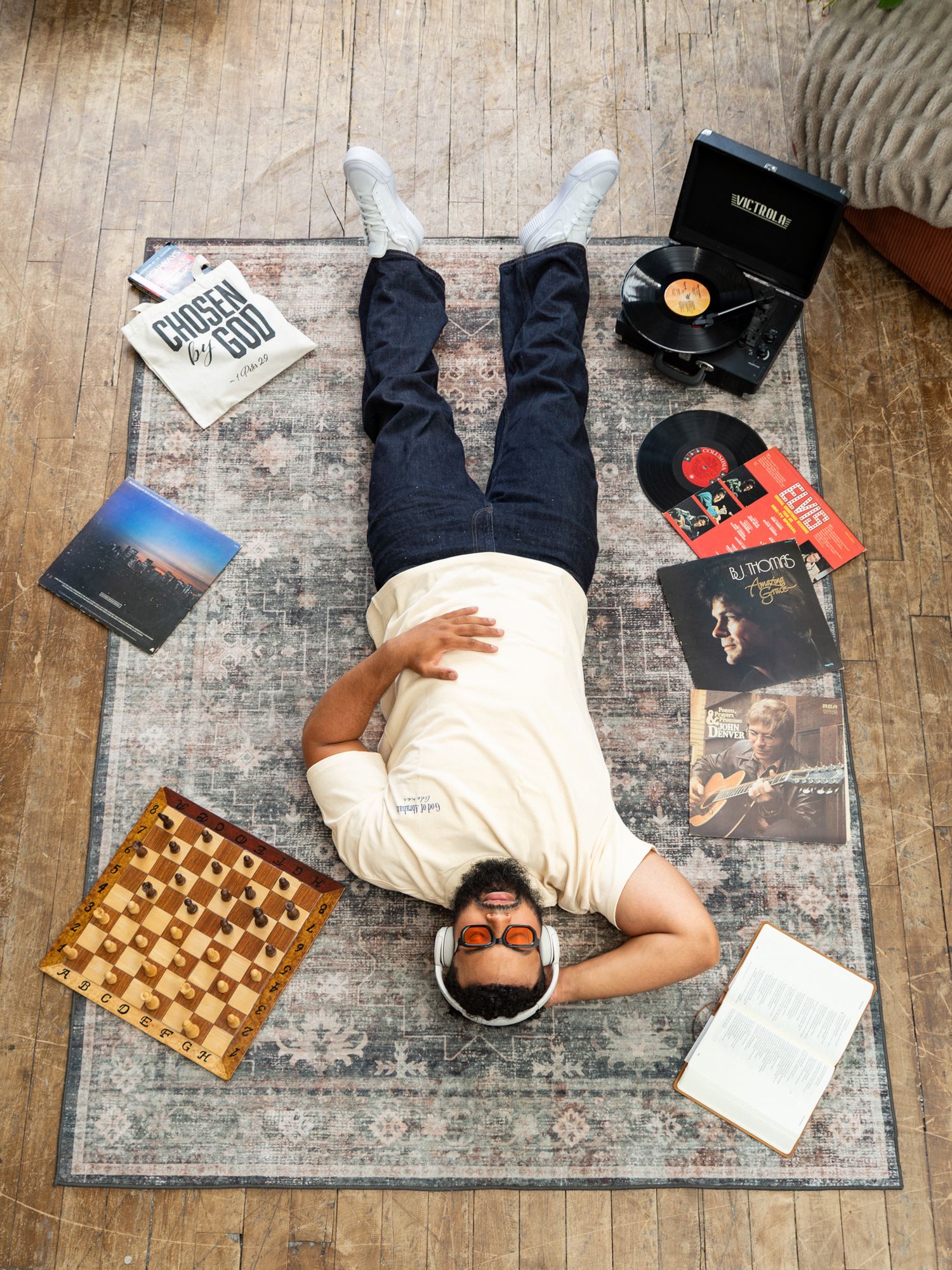 Man lying on rug surrounded by vinyl records, chess board, headphones, and books on wooden floor.
