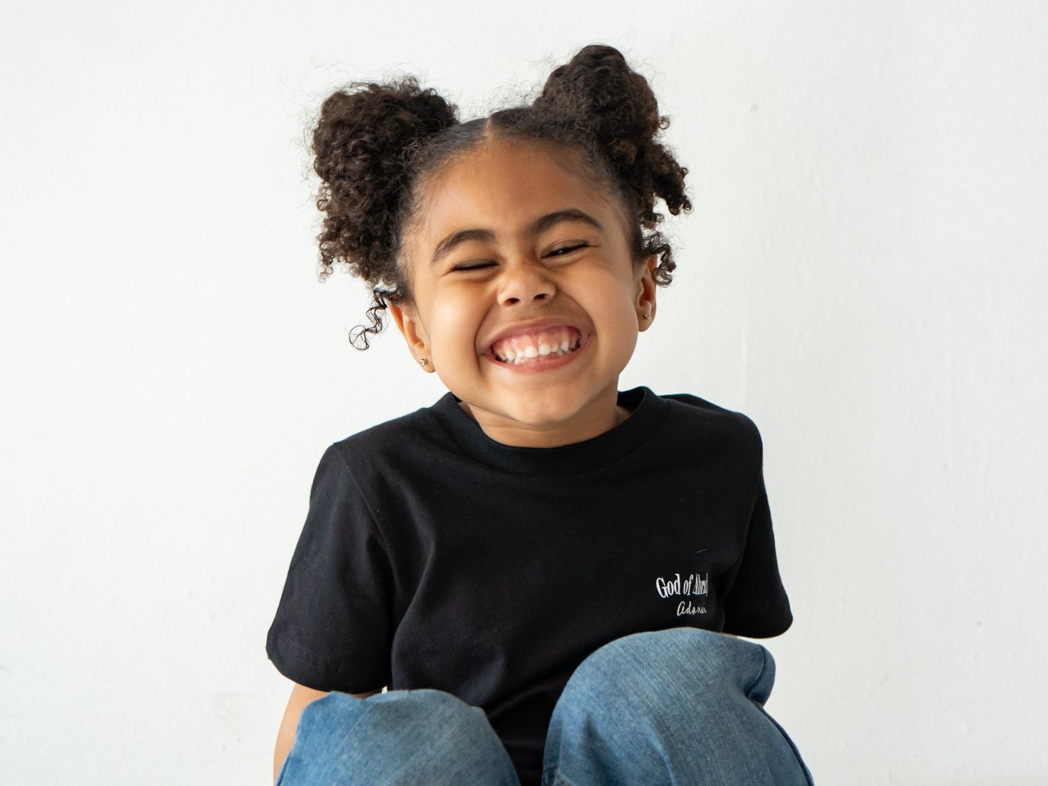 Joyful young girl with curly puff hairstyle laughing in black graphic tee and jeans on white background.