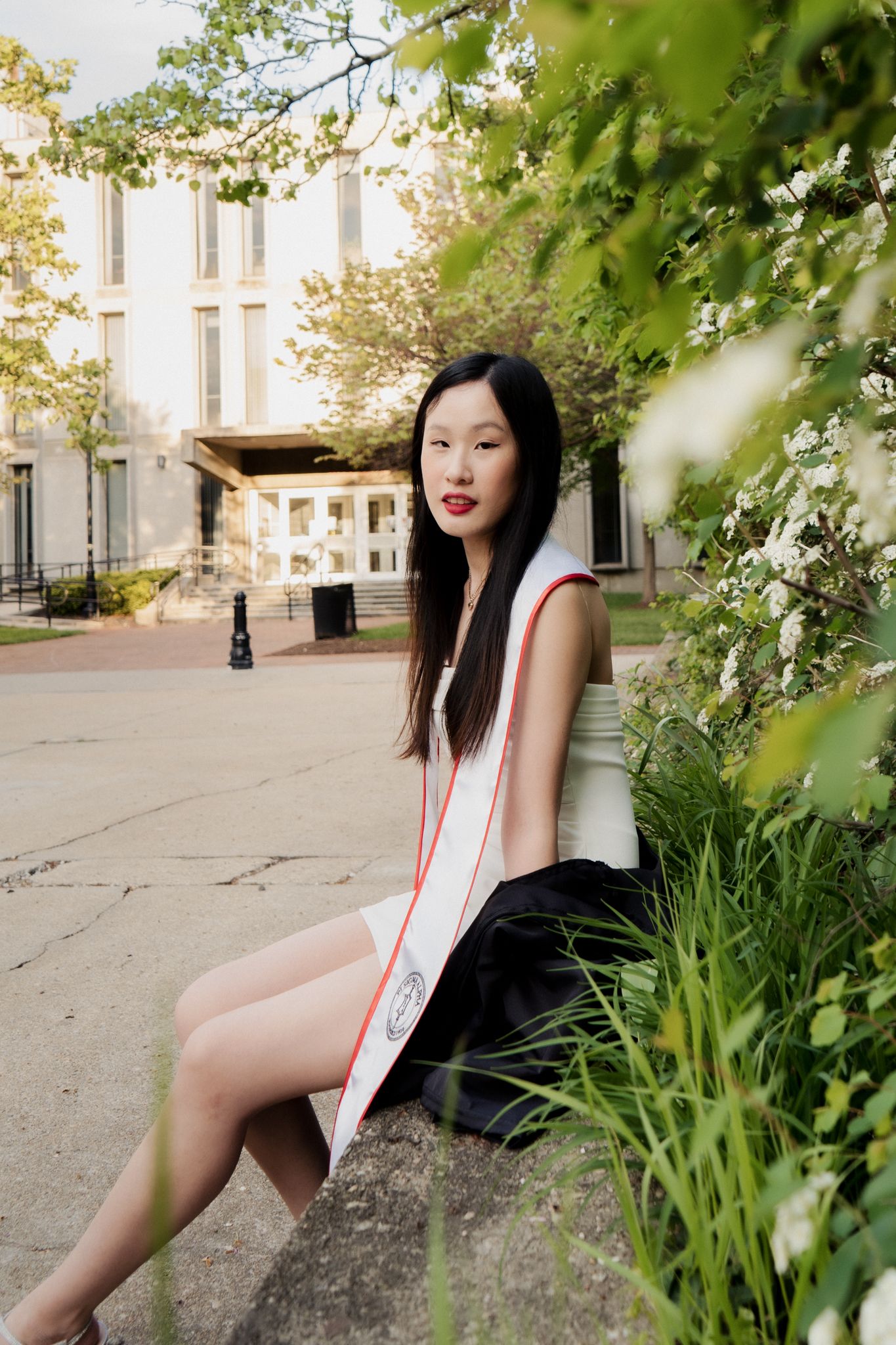 Young Asian woman in graduation sash sits on stone ledge outdoors near blooming flowers and campus building.