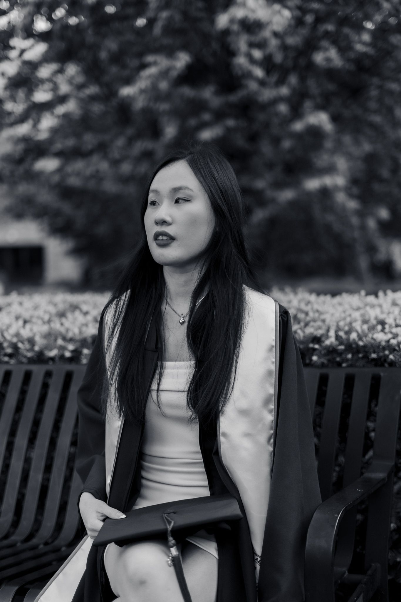 Black and white portrait of a young Asian woman in graduation gown sitting on a park bench.