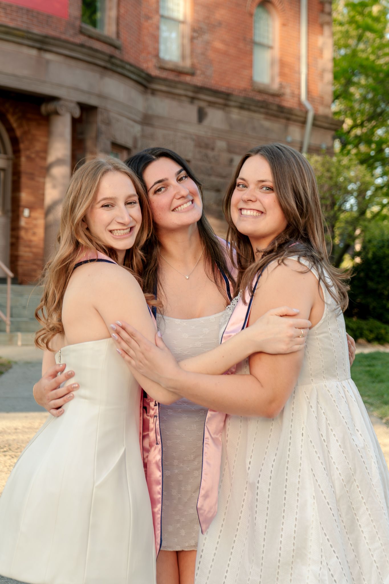 Three friends in light colored dresses embrace while posing against brick building.