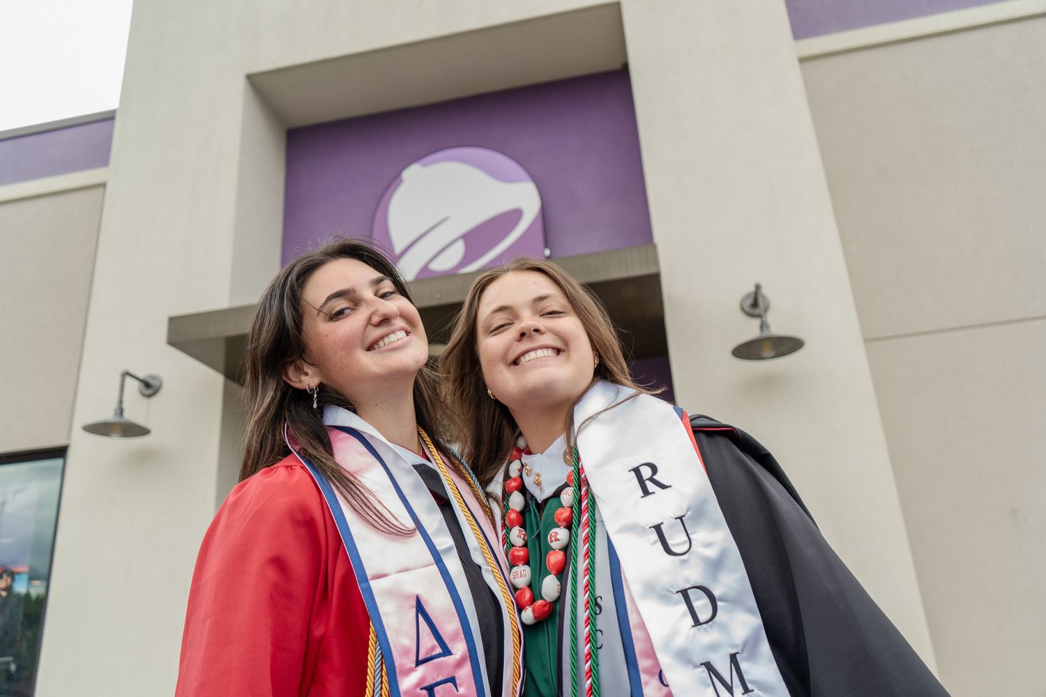 Graduates wearing colorful stoles and sashes pose together outside a building with purple accents.