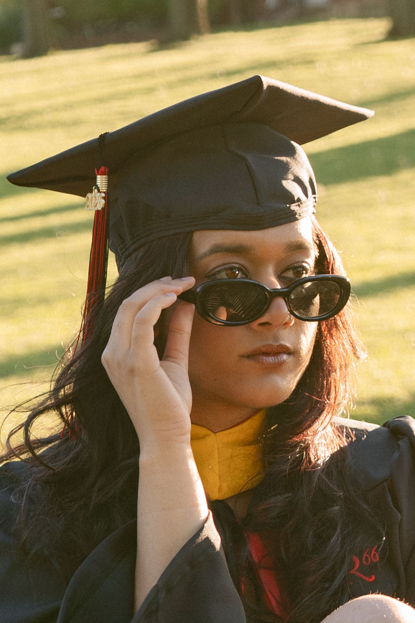 Young woman in black graduation cap and gown adjusts sunglasses outdoors on green lawn.