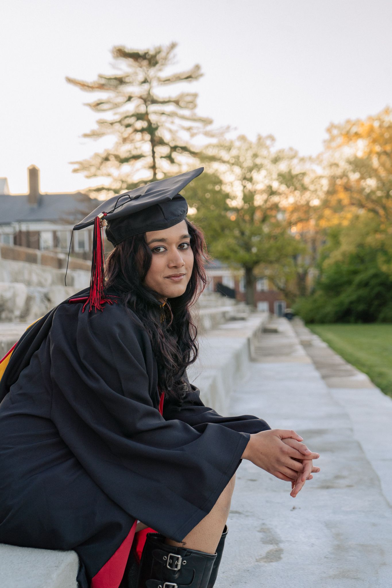 Young woman in black graduation cap and gown sitting outdoors on steps, smiling at camera on a sunny day.