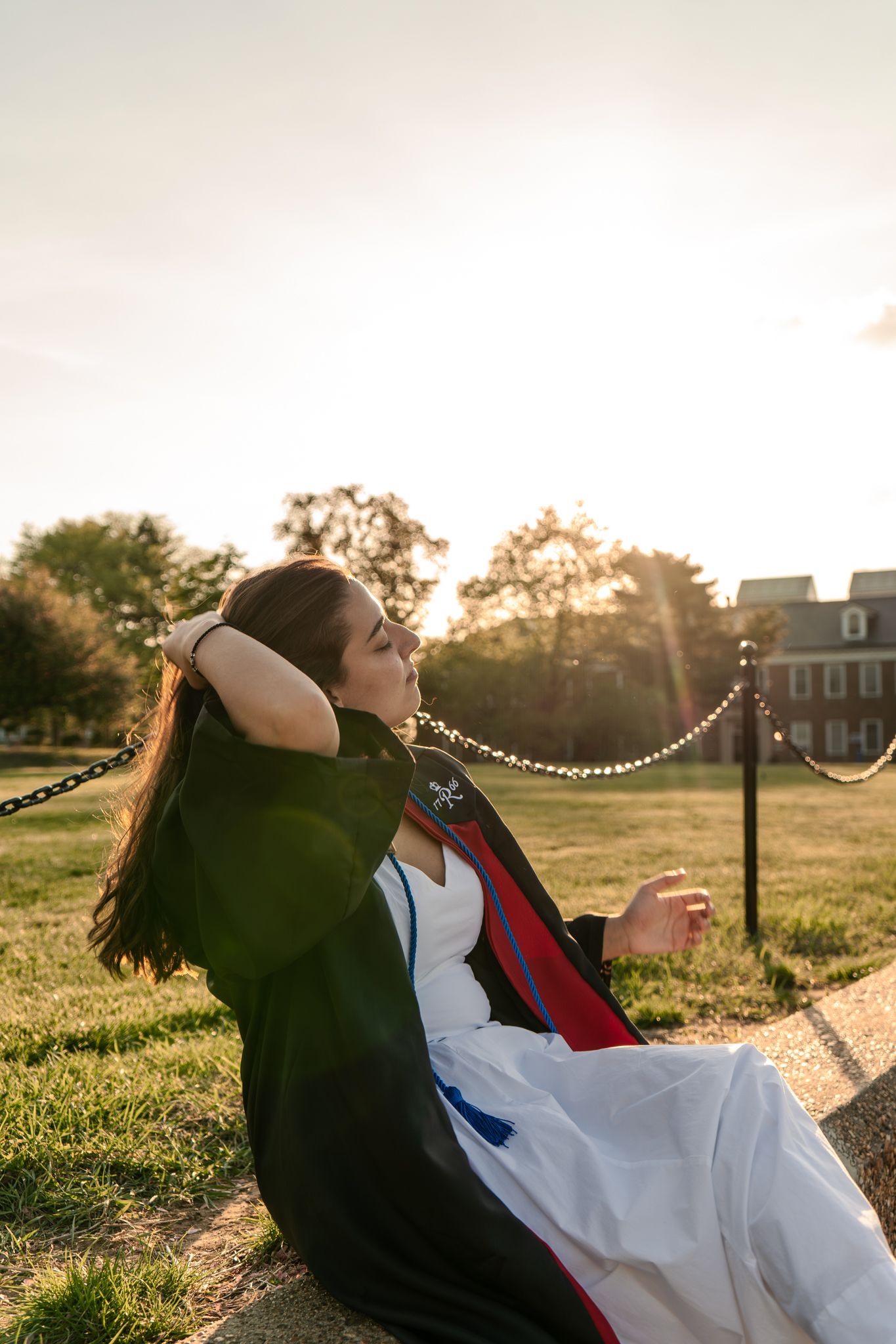 Graduate in academic regalia relaxes on grass during golden hour photoshoot.