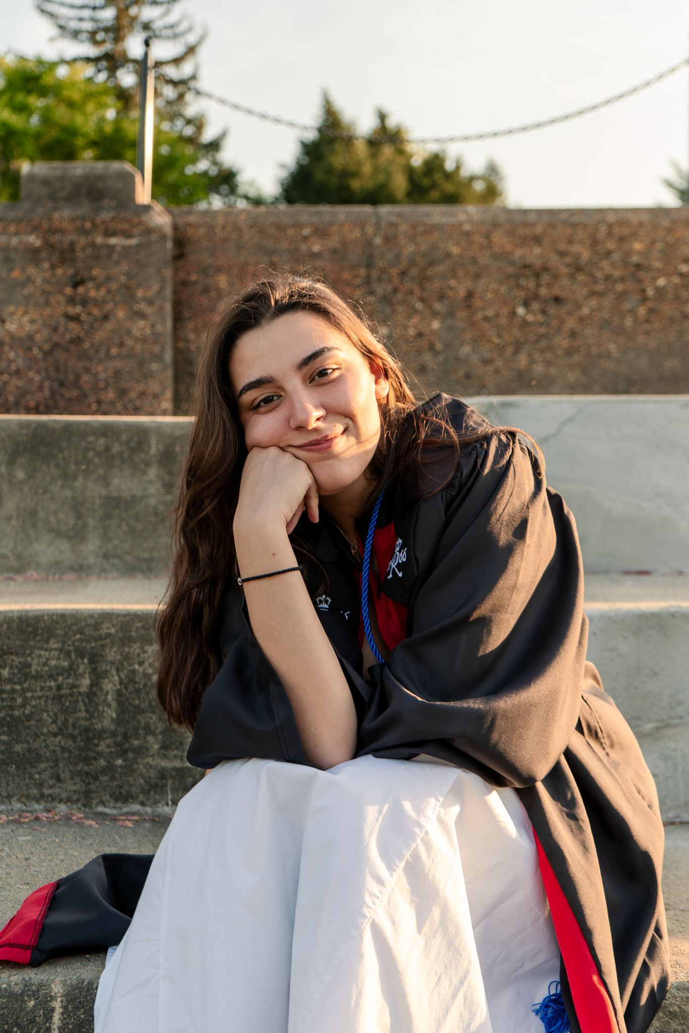 Graduate in black academic gown poses against concrete wall during sunset.