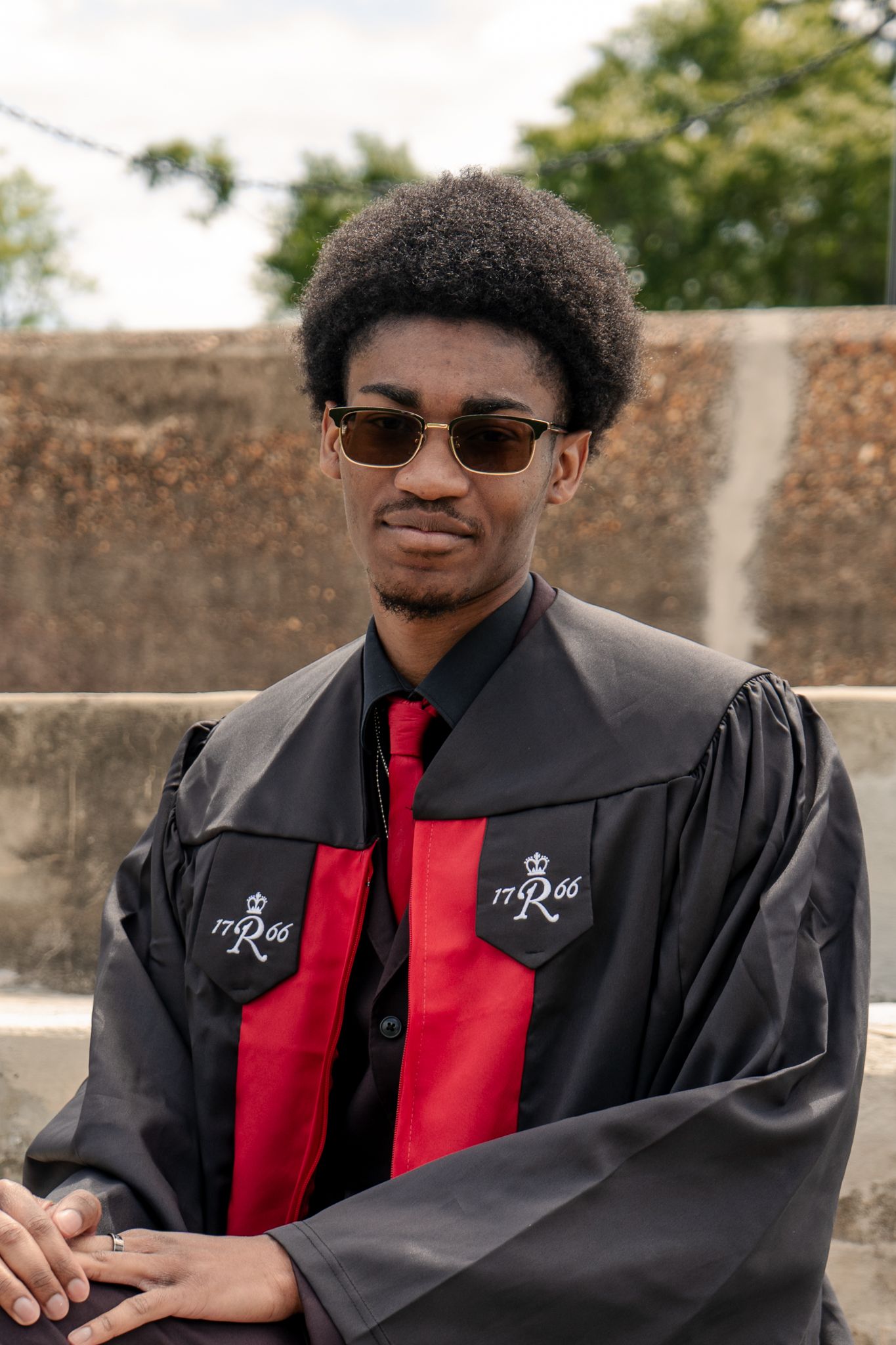 Graduate in black and red regalia poses against stone wall with sunglasses.
