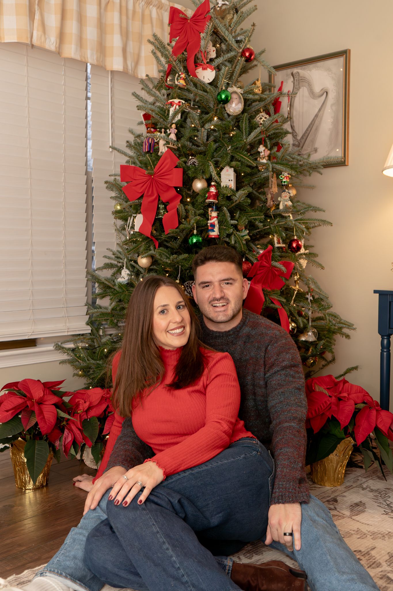 A couple in red and gray sits together in front of a decorated Christmas tree with red bows and ornaments.