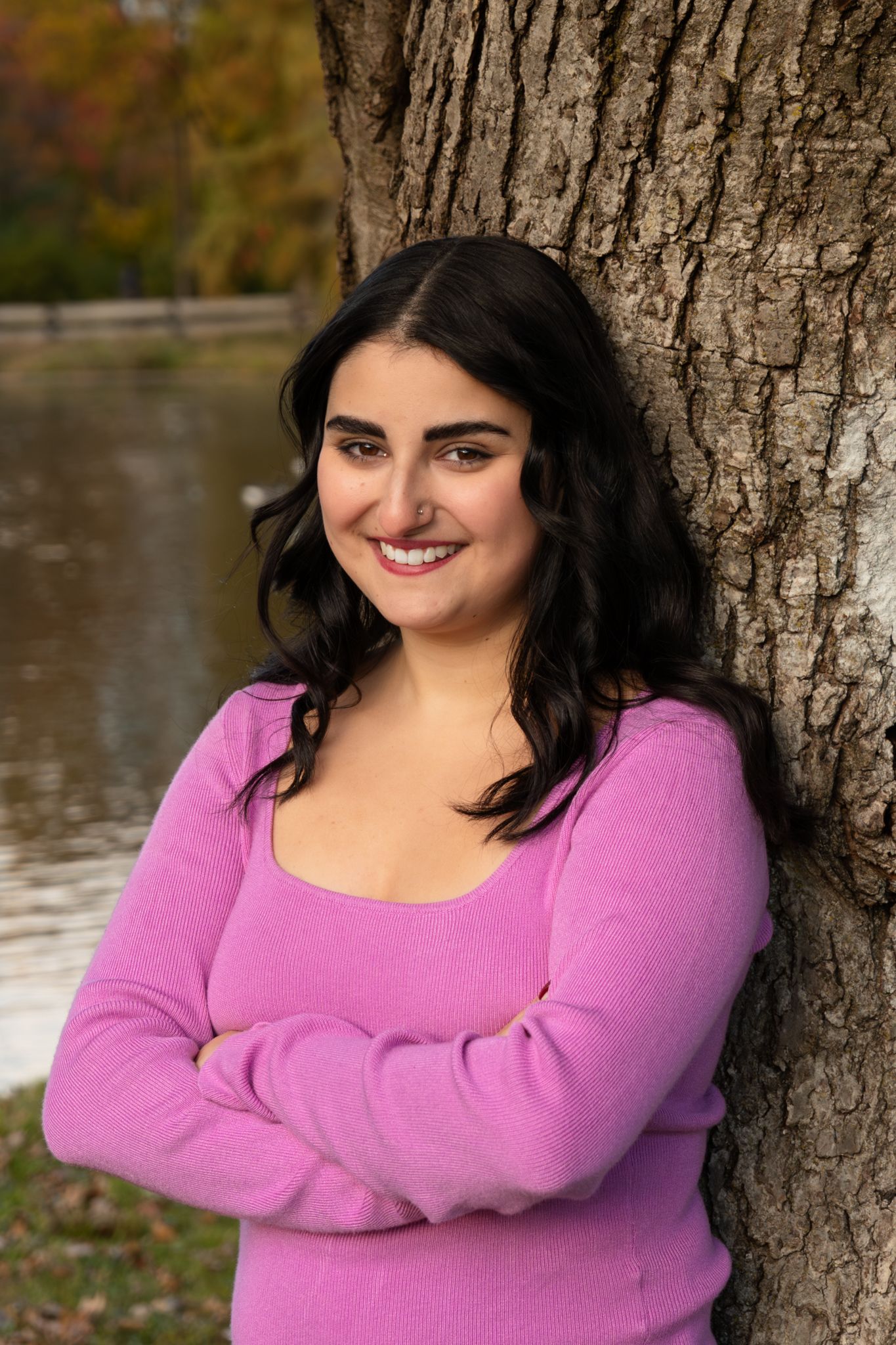 Person in pink sweater poses against tree trunk by water in autumn outdoor portrait series.