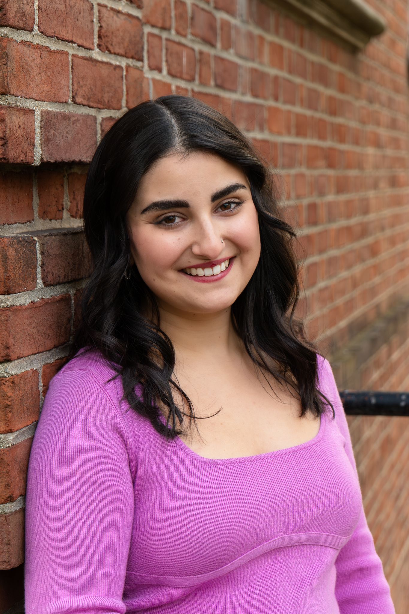 A person in a bright magenta sweater poses against a brick wall while smiling warmly at the camera.