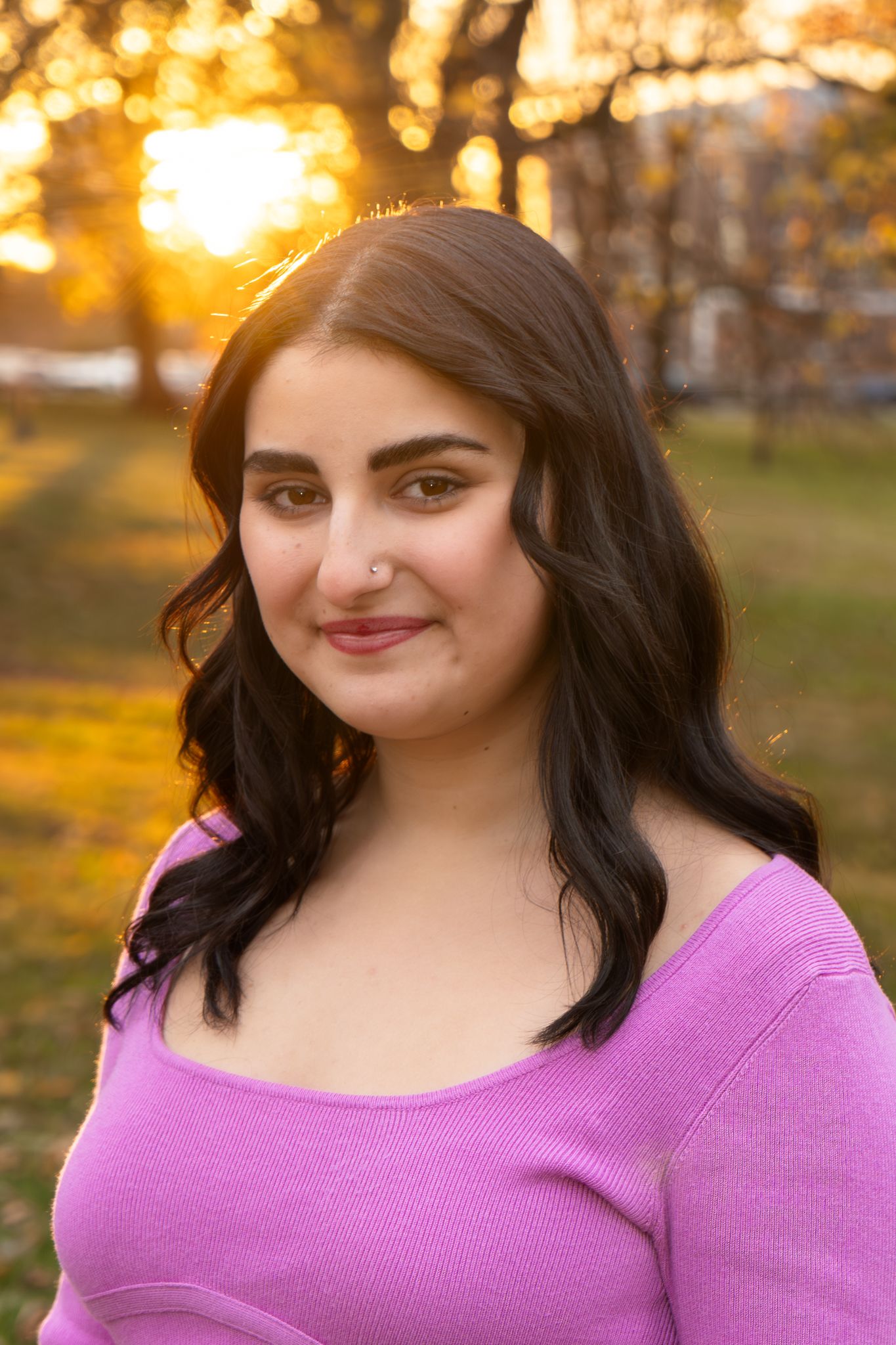 A woman in a bright purple sweater poses outdoors during golden hour with autumn trees in the background.