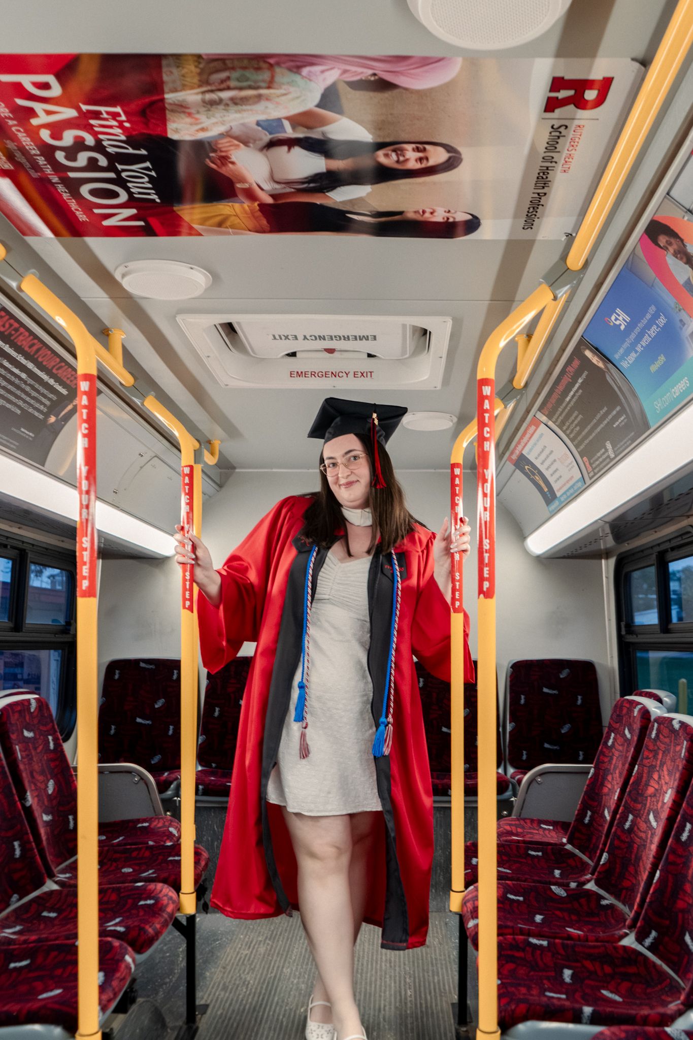 Graduate in red academic regalia poses playfully inside a bus with maroon seats.