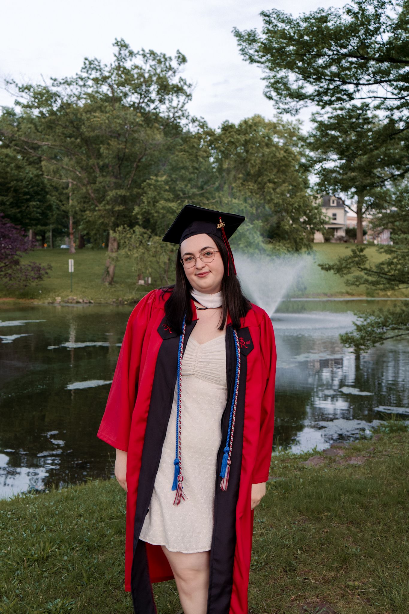 Graduate in red regalia poses near a scenic pond with fountain on a sunny day.