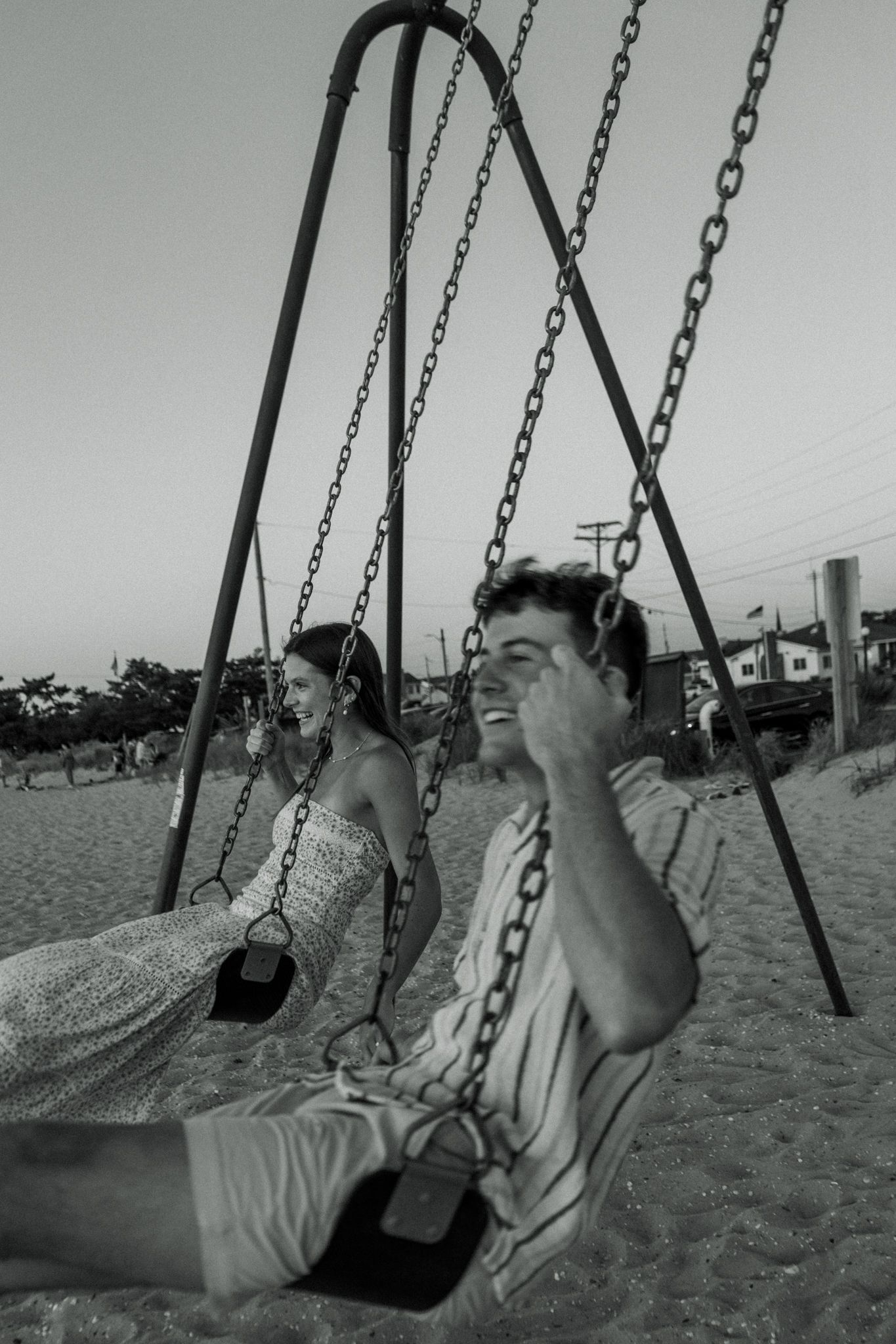 A playful black and white photo of a couple on swings at the beach.