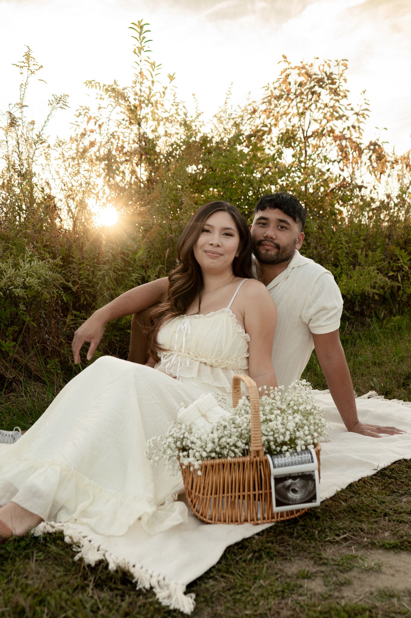 A couple sits on a blanket at sunset with a wicker basket of baby's breath flowers and ultrasound photo nearby.