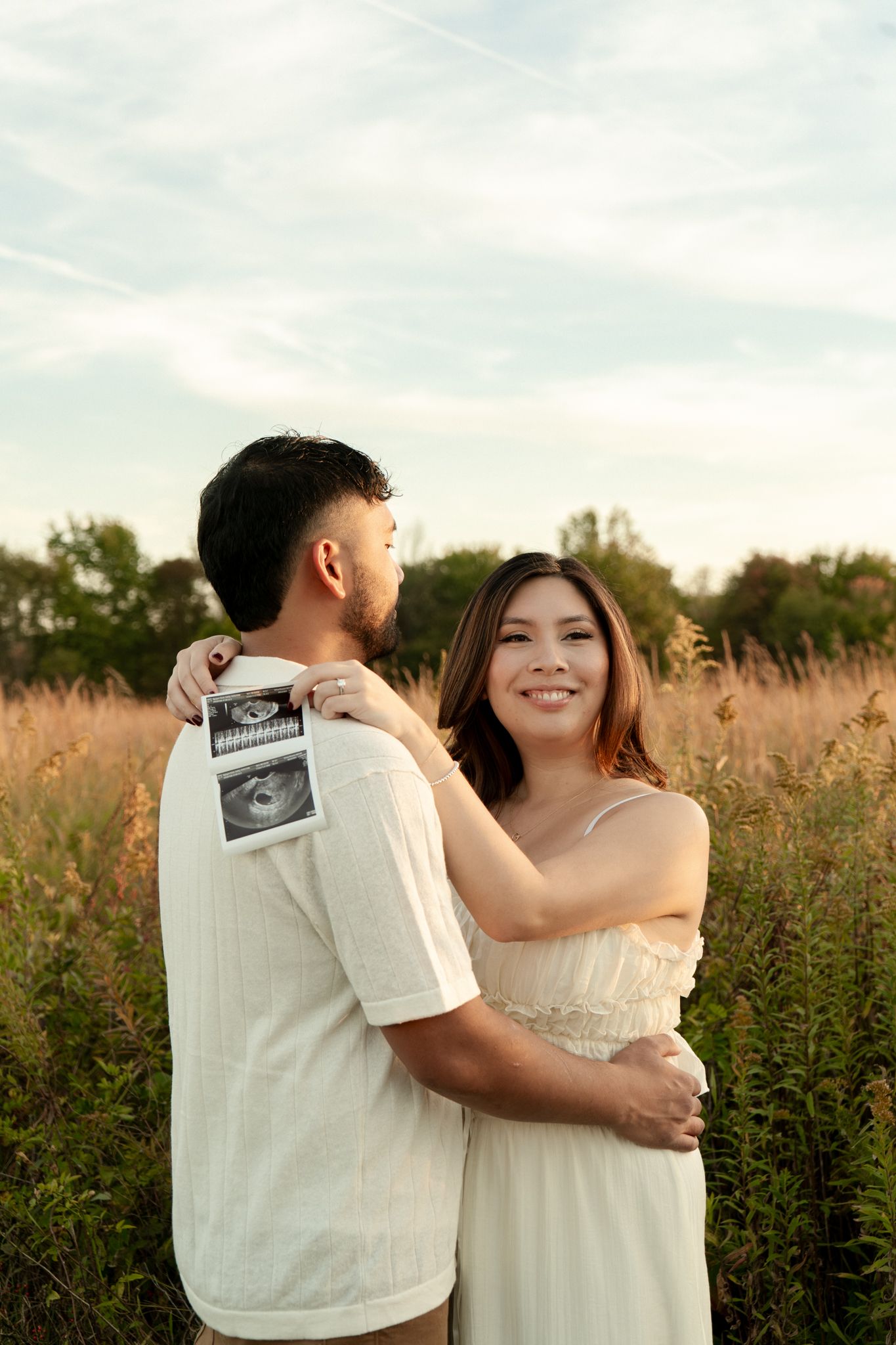 A couple in a wildflower field holds up ultrasound photos during a pregnancy announcement photoshoot at sunset.