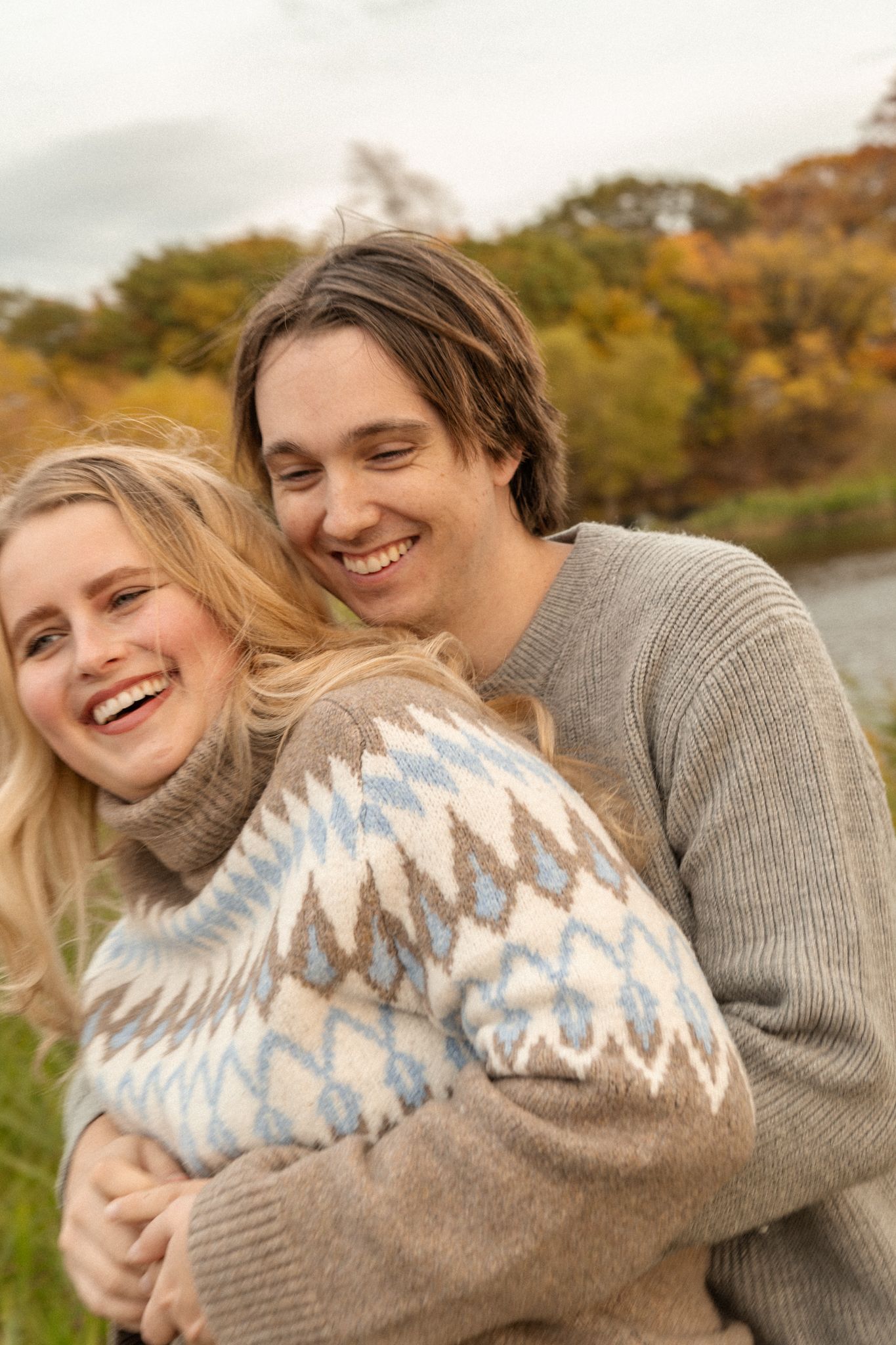 Couple wearing cozy sweaters embrace and laugh together in an autumn outdoor setting.