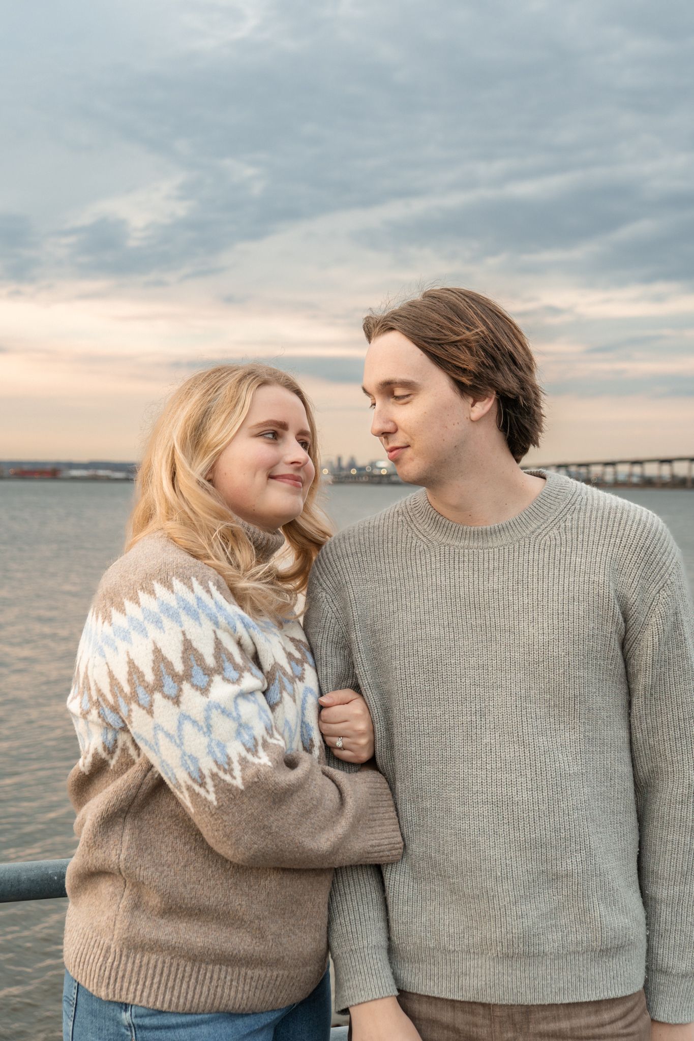 A couple in matching gray sweaters embrace and smile while standing by the ocean at sunset.