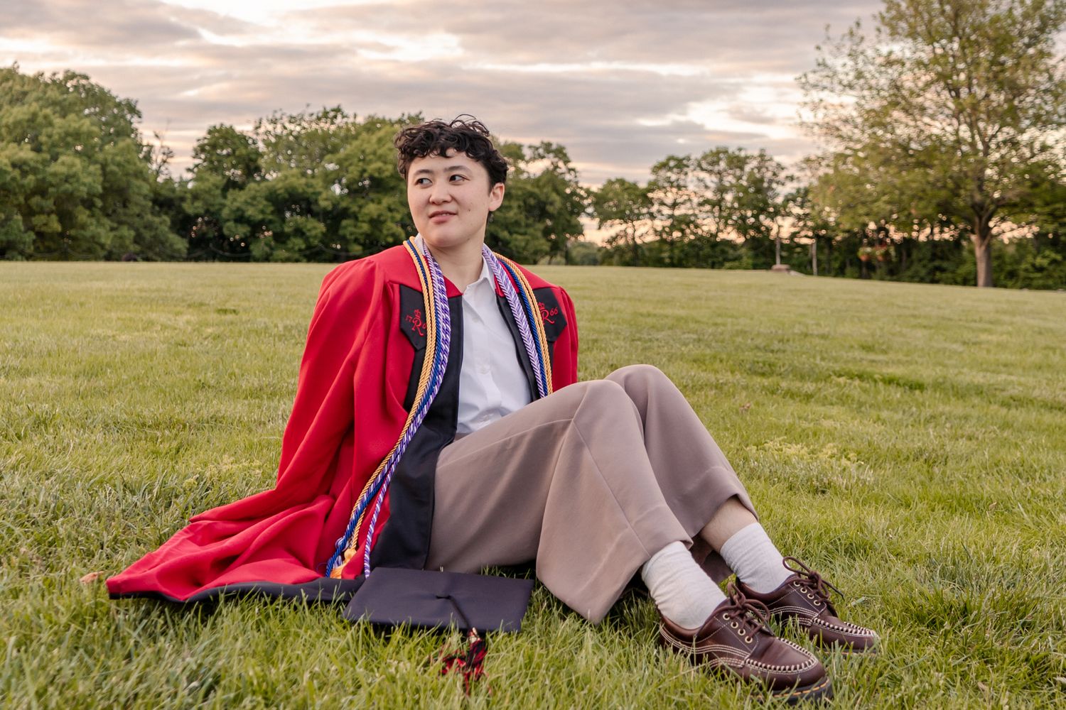 Graduate in red regalia relaxing on grassy hill during sunset.