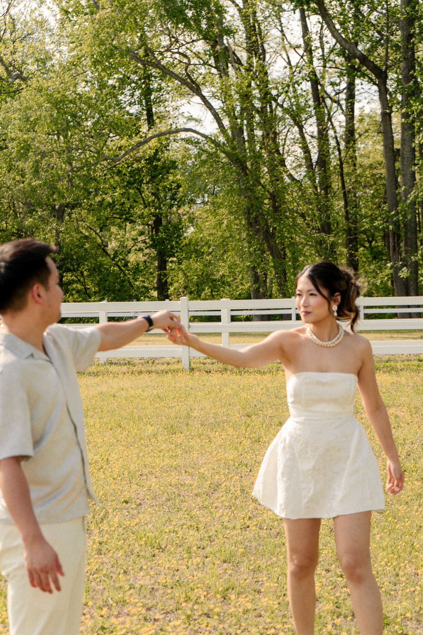 Couple in white attire dance together in a sunny field with white fence in background.