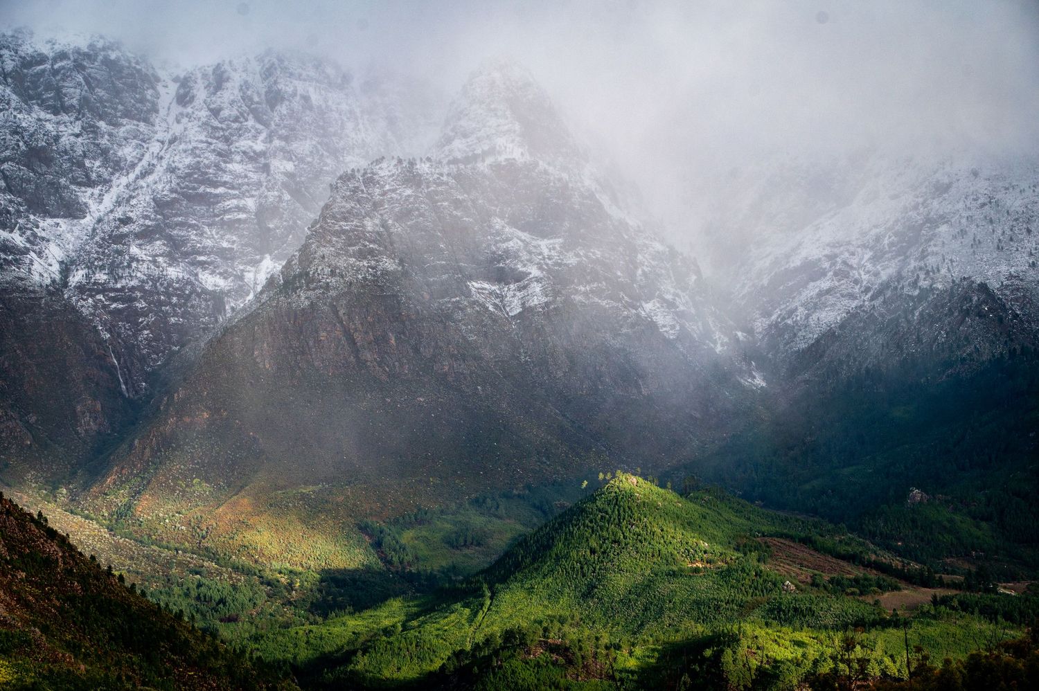 Dramatic mountain landscape with snow-capped peaks and sunlit green valley below in misty conditions.