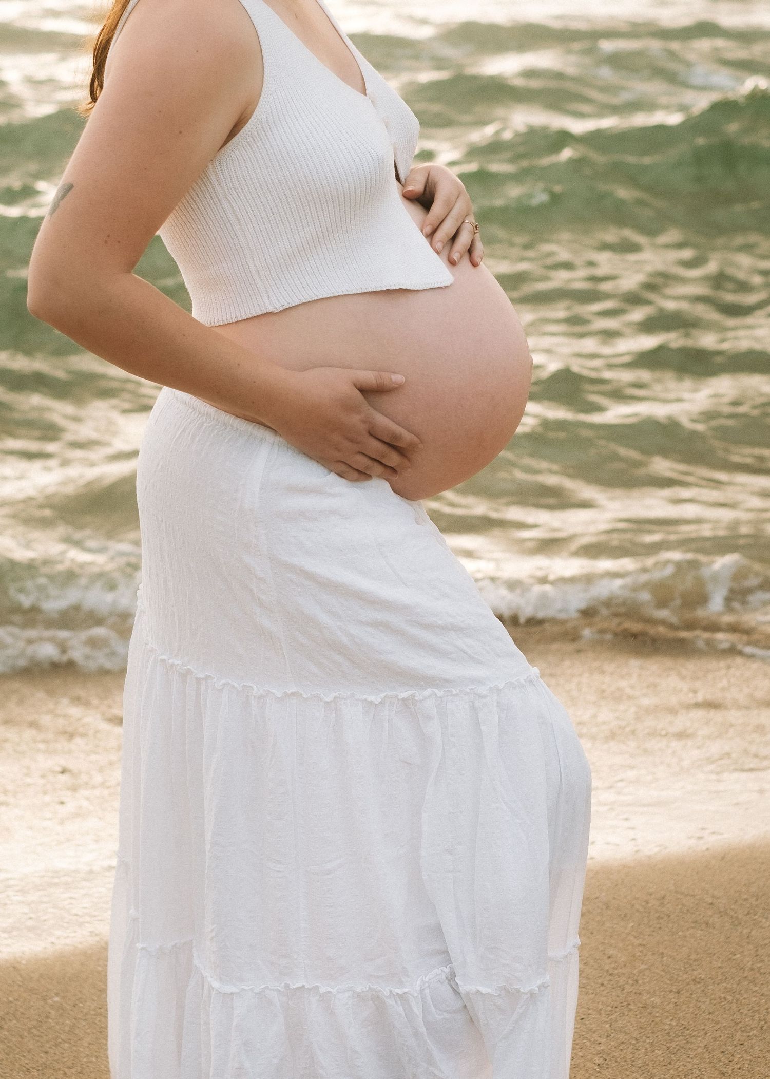 Pregnant woman in white dress stands on beach with ocean waves in background during sunset.